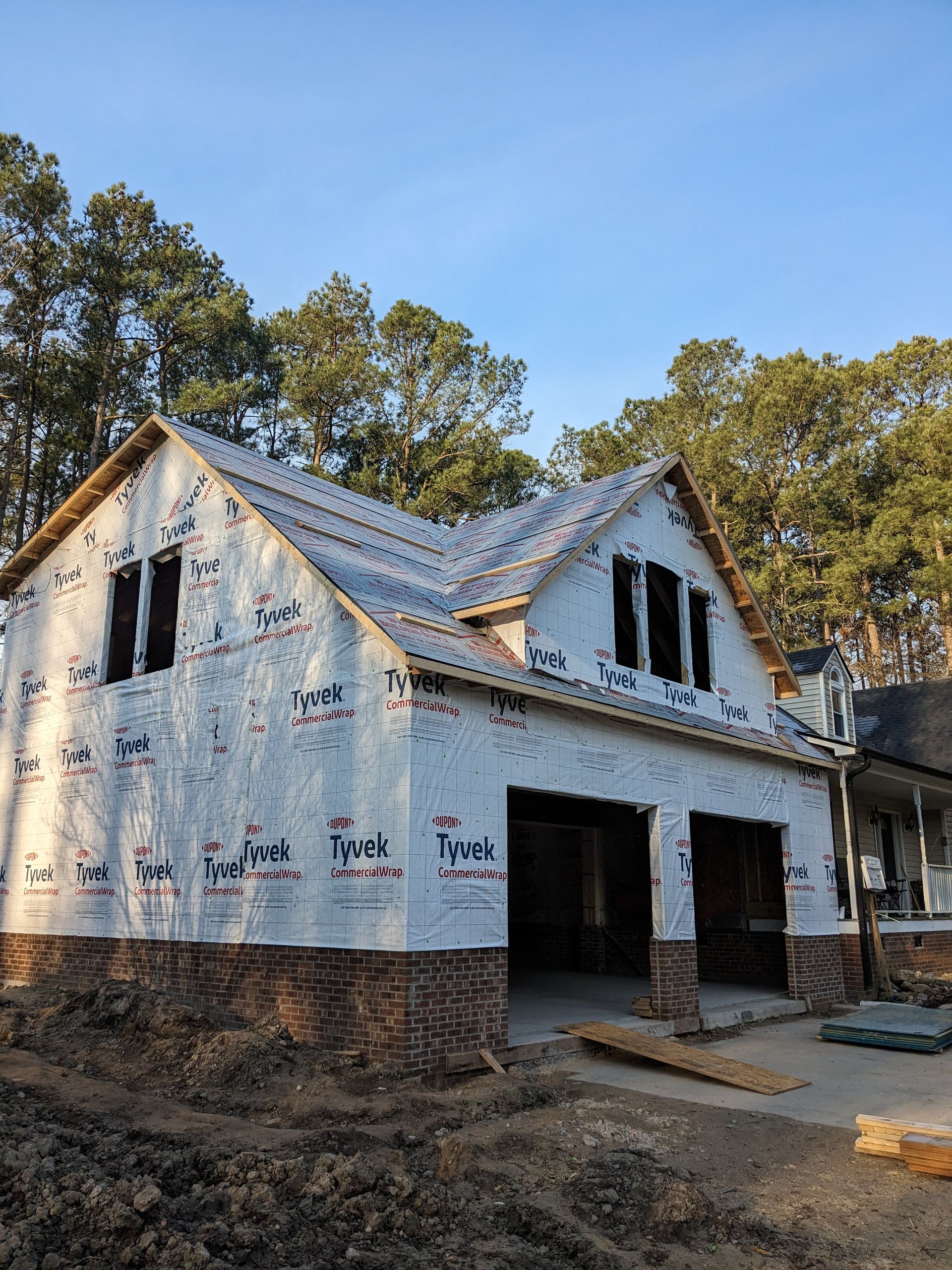 A partially constructed two-story house with a brick base and Tyvek-covered exterior walls in a wooded setting.