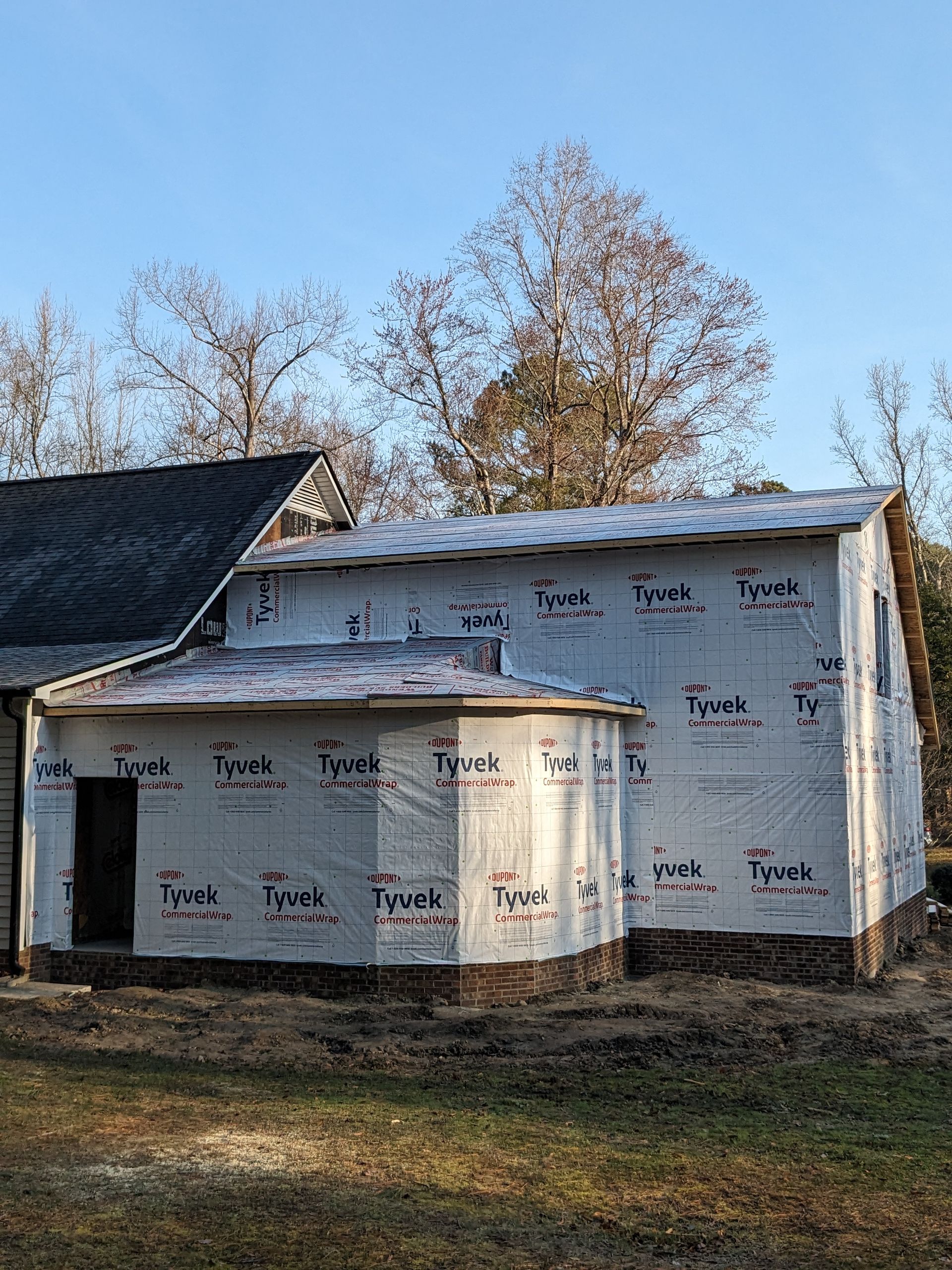 A house addition under construction, covered in white Tyvek wrap with a stone veneer base, set against bare trees.