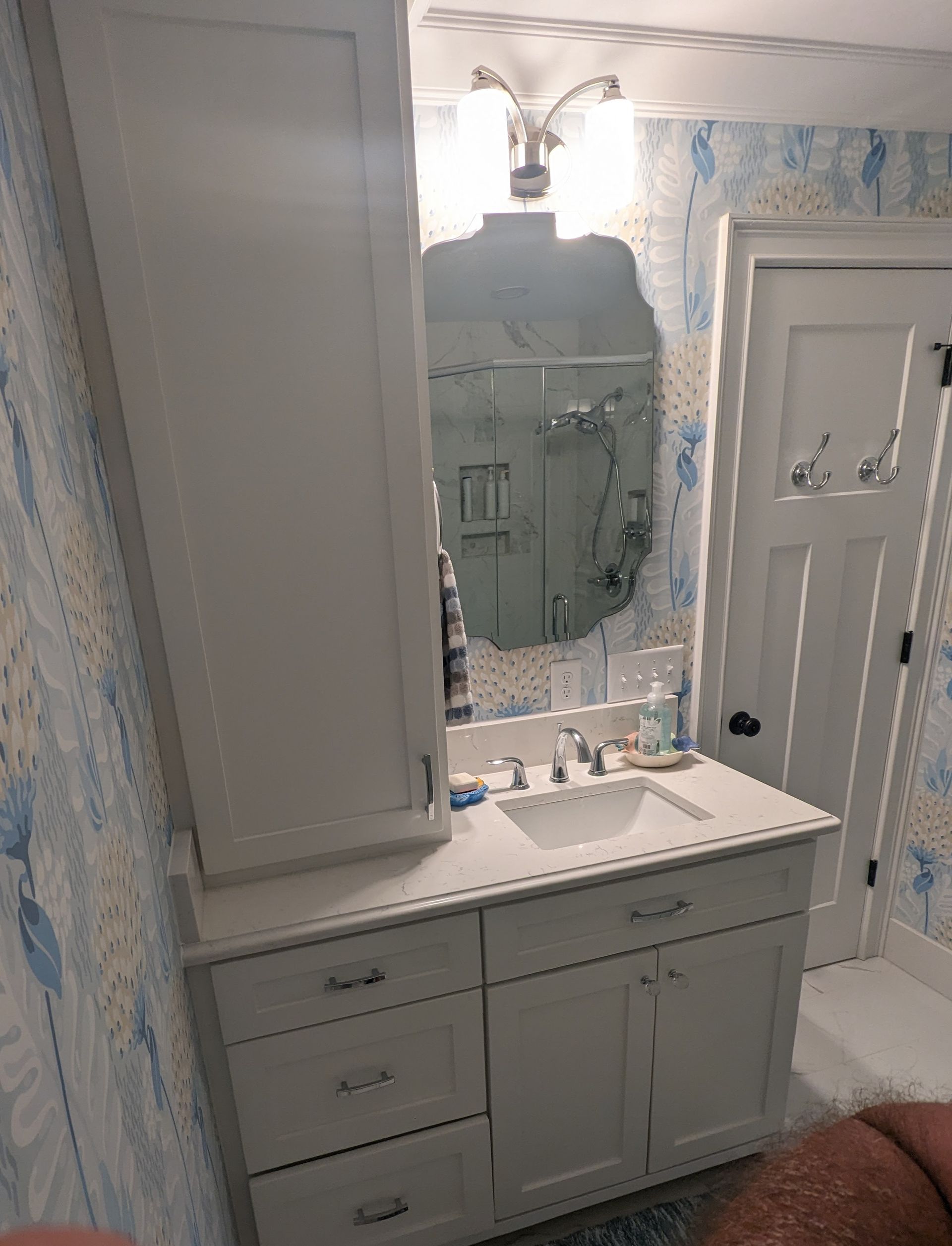 A gray bathroom vanity with a tall side cabinet, white countertop, sink, mirror, and light fixture against patterned wallpaper.