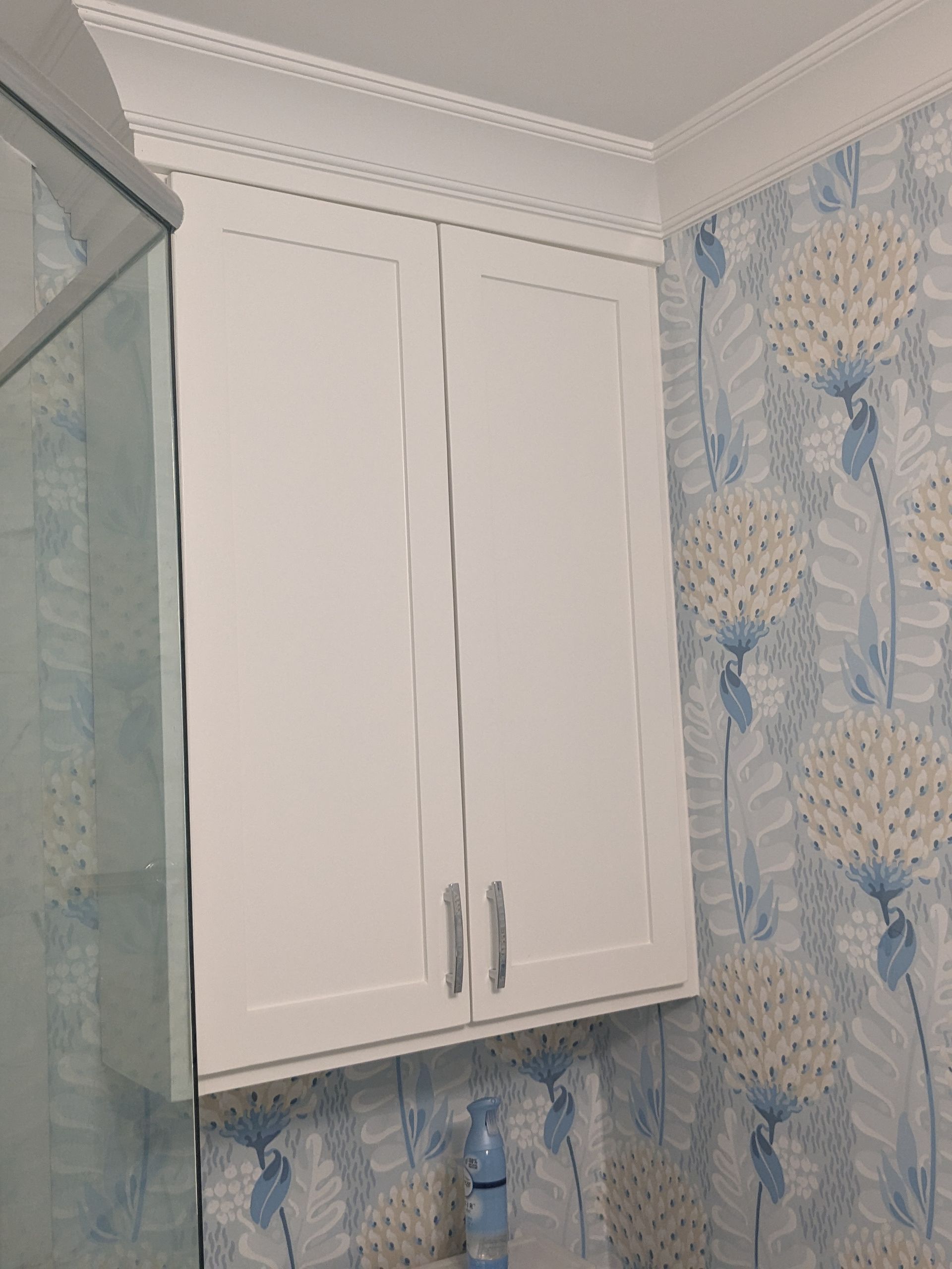 White shaker-style wall cabinets installed against light blue and cream floral patterned wallpaper in a bathroom.