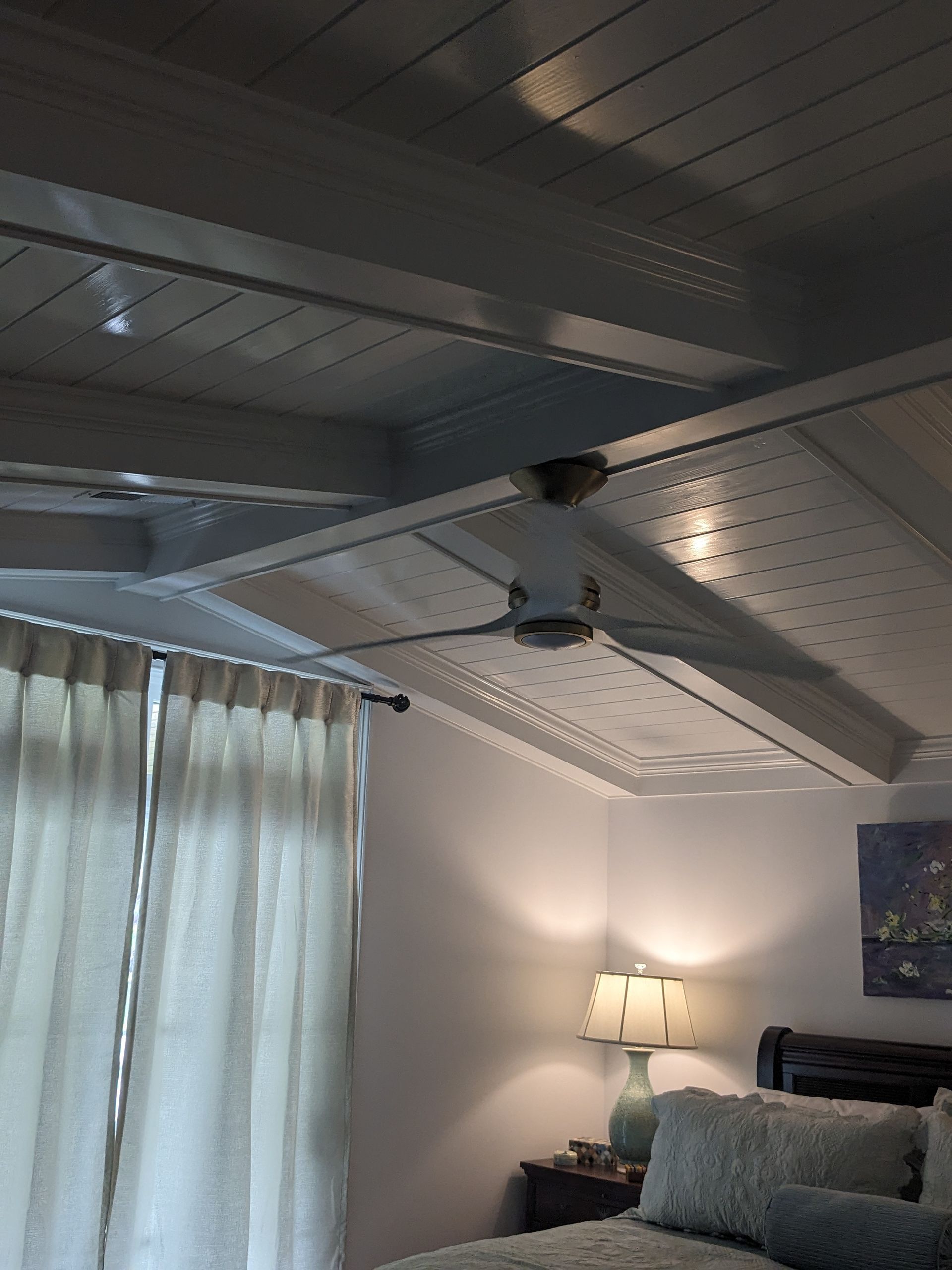 A bedroom ceiling with white wooden beams and a white ceiling fan, alongside beige curtains and a lamp on a nightstand.