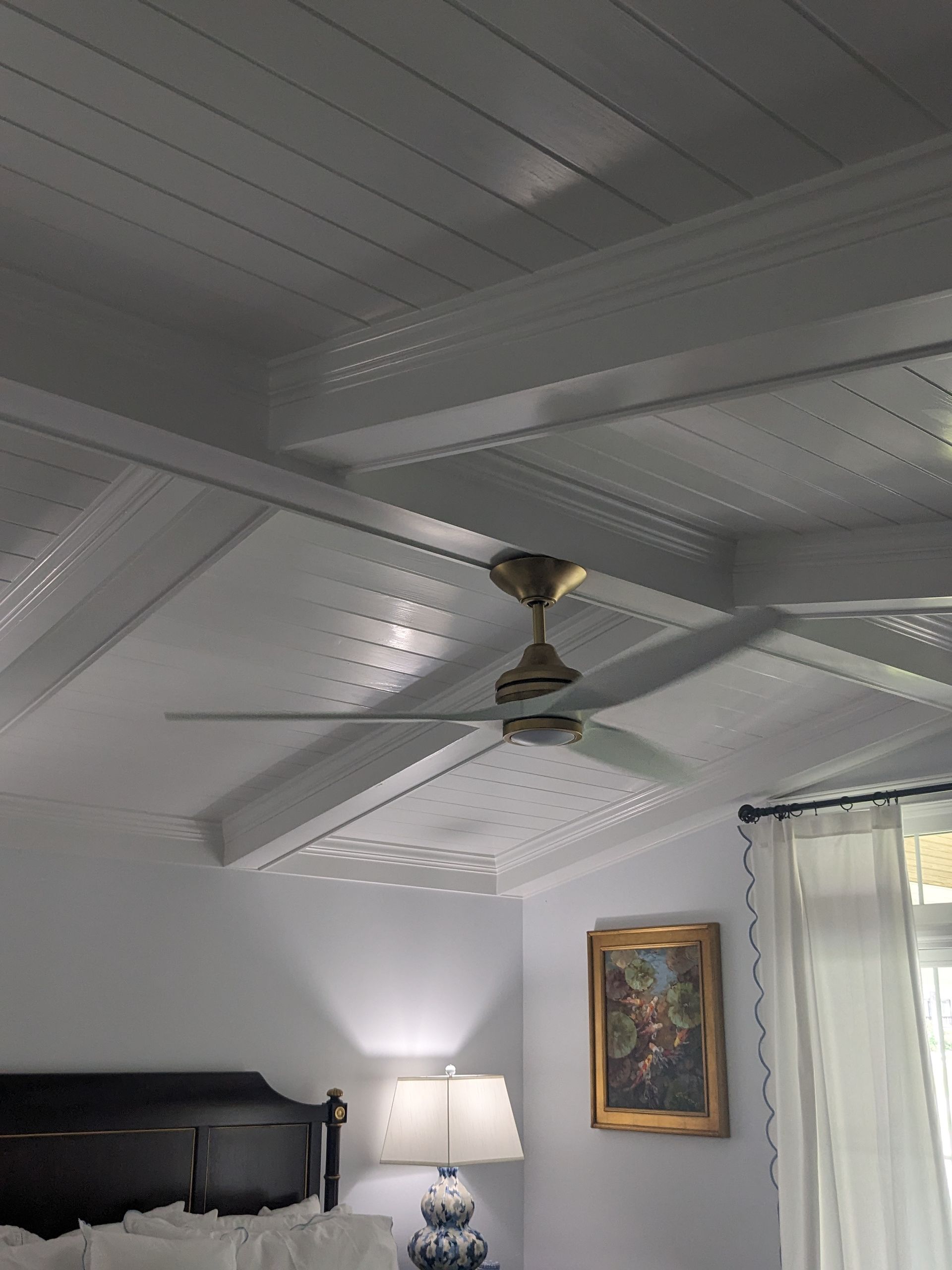 A ceiling fan hangs from a white paneled ceiling above a bedroom with a framed painting and a bedside lamp.