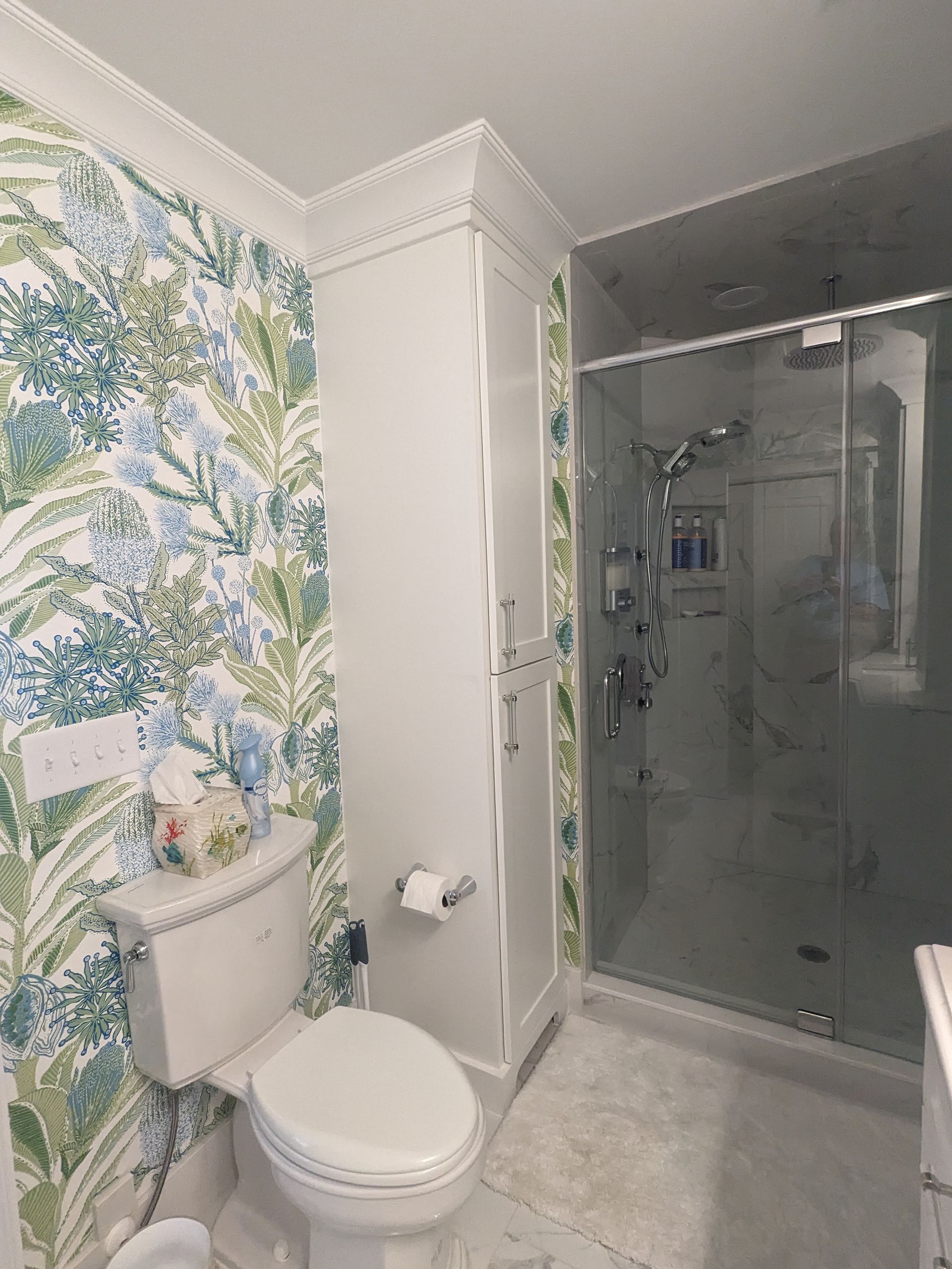 A bathroom with blue and green botanical wallpaper, a white toilet, a tall storage cabinet, and a glass-enclosed shower.