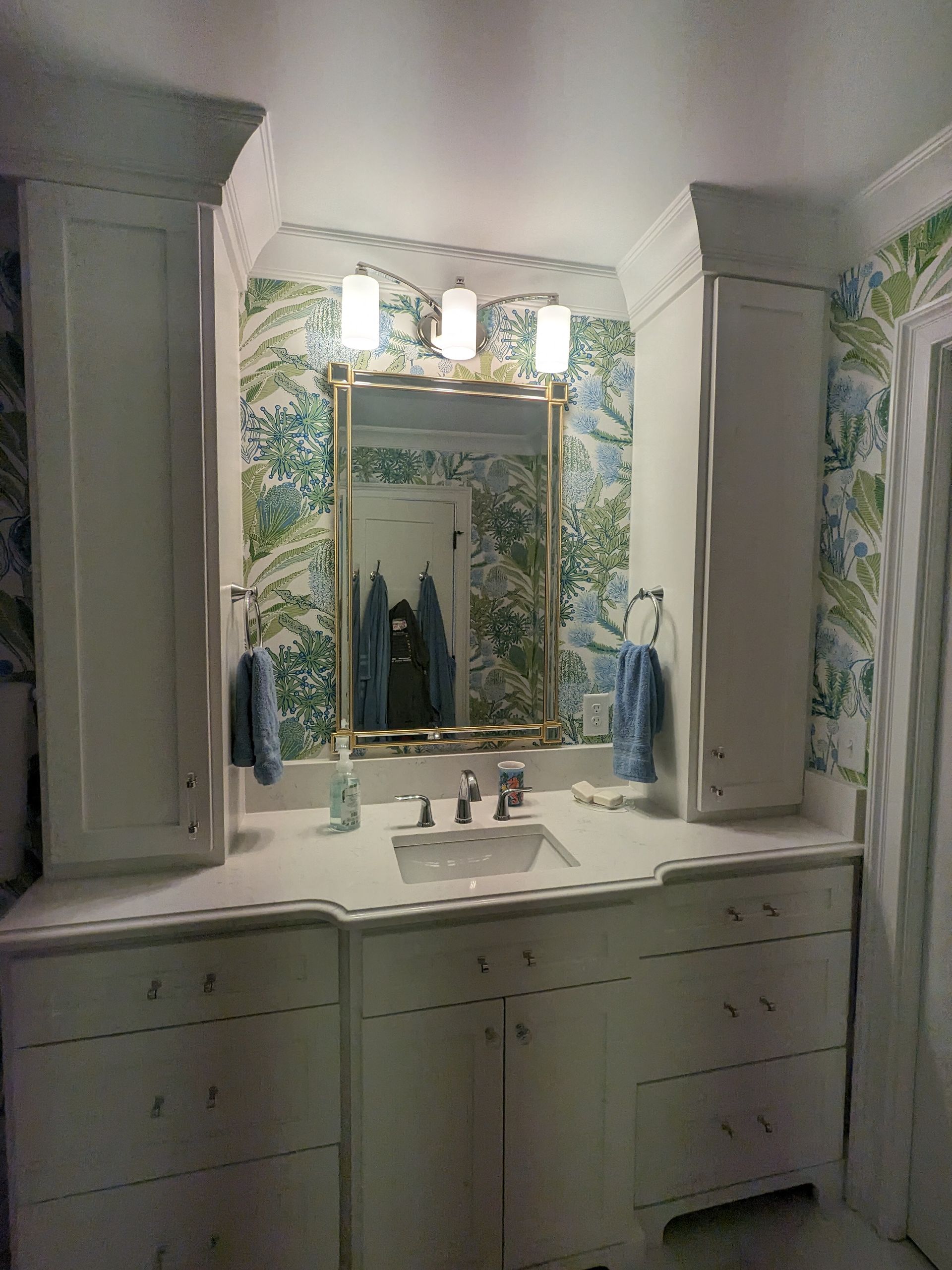 A bathroom vanity with white cabinets, a central sink, an ornate gold mirror, and vibrant blue and green botanical wallpaper.