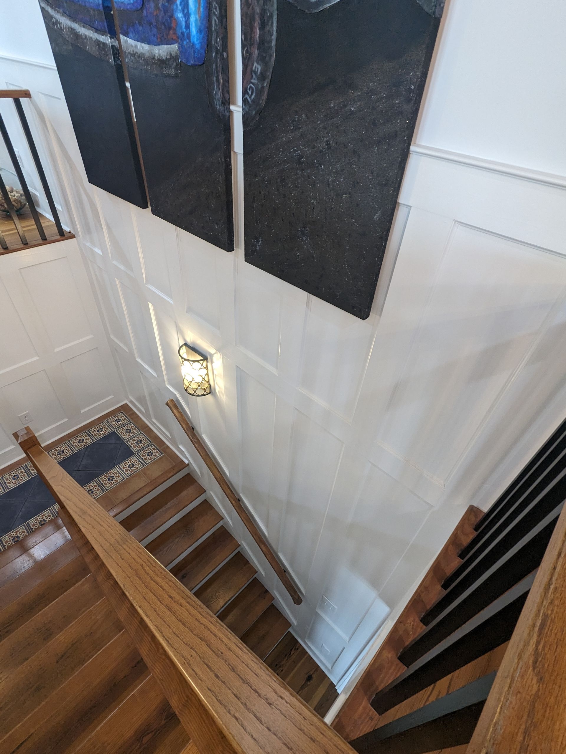 A downward view of a wooden staircase with a wall-mounted light fixture, decorative wall paneling, and art panels above.