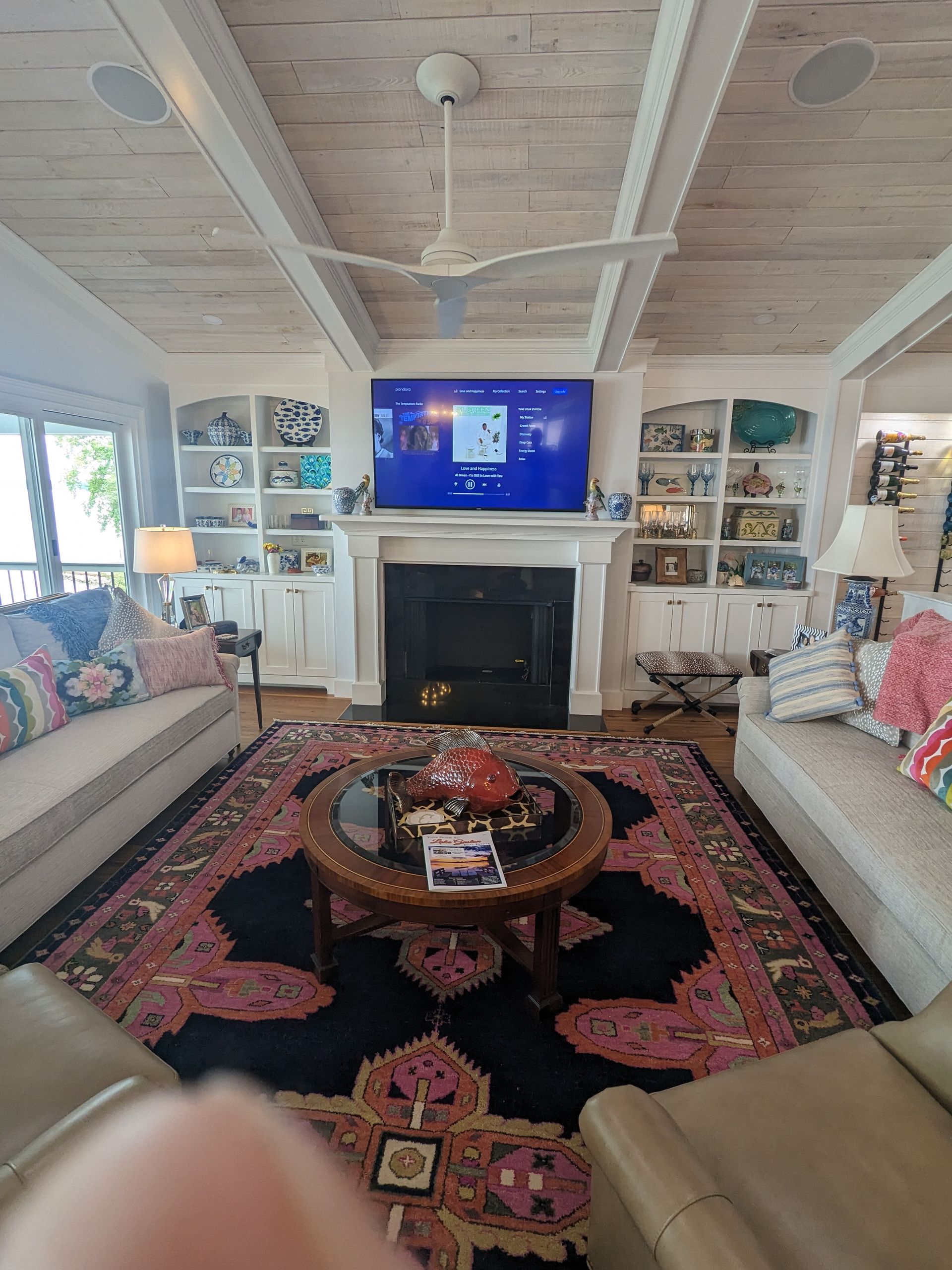 A cozy living room with a black fireplace, a large TV, built-in shelves, and a patterned rug on a white wood ceiling room.