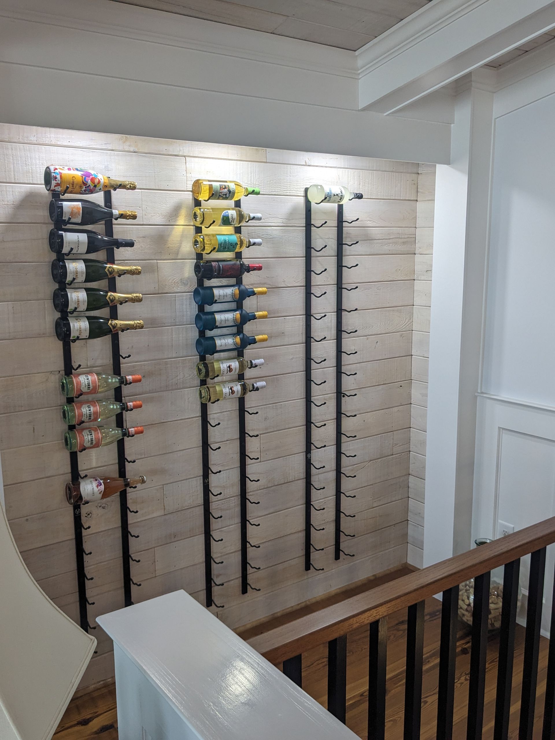Wall-mounted wine racks holding bottles on a light-colored wood slat wall, positioned above a stairwell railing.