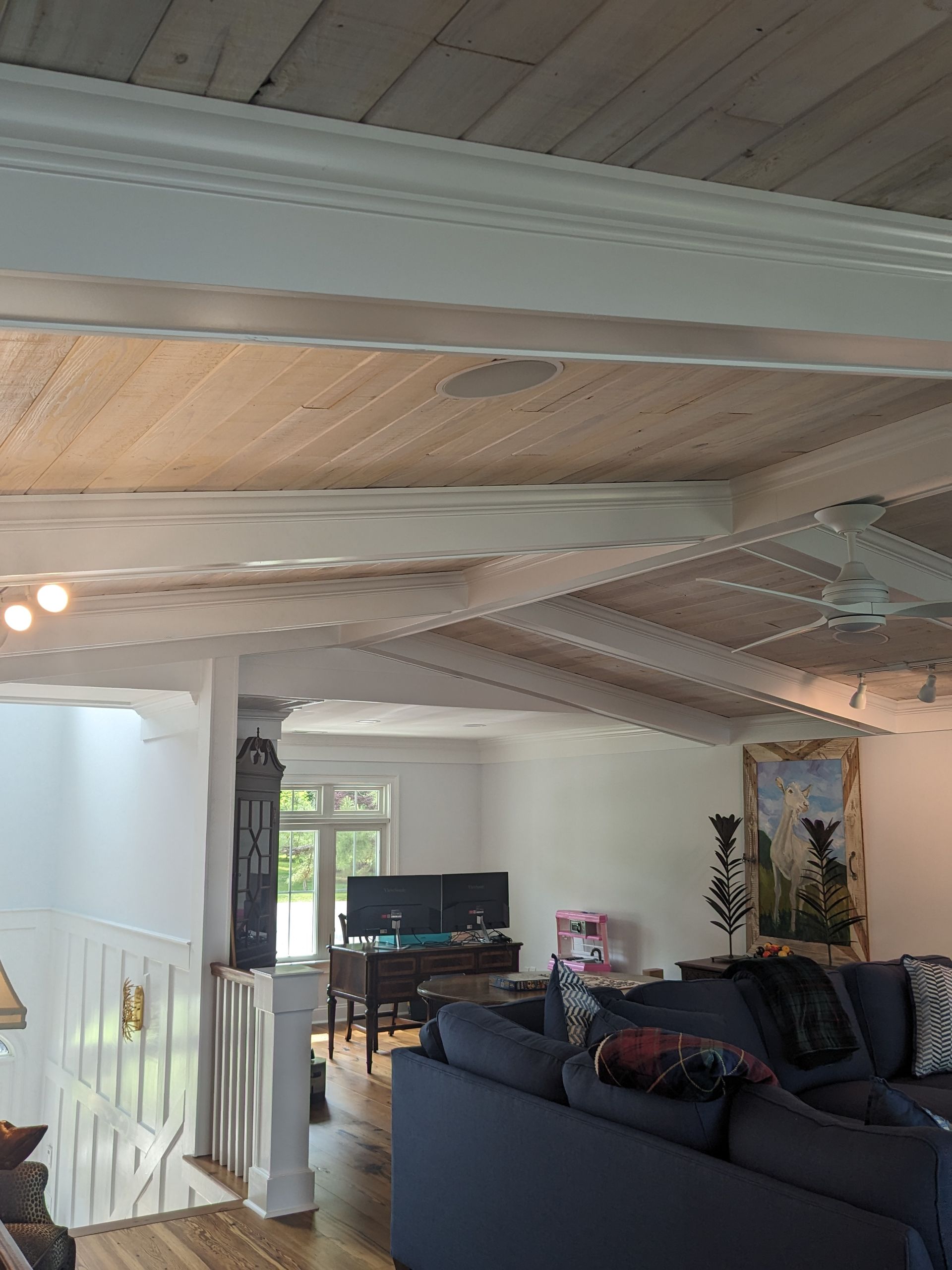 A cozy living room with a wood-planked ceiling, white exposed beams, a dark blue sofa, and a view into a bright office area.
