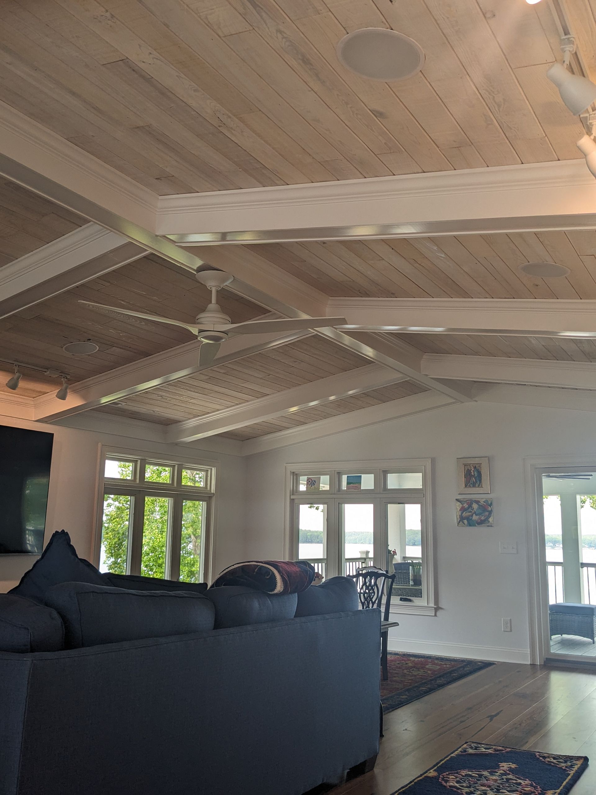 A living room with a white planked vaulted ceiling, wood beams, a ceiling fan, a dark sofa, and windows overlooking water.