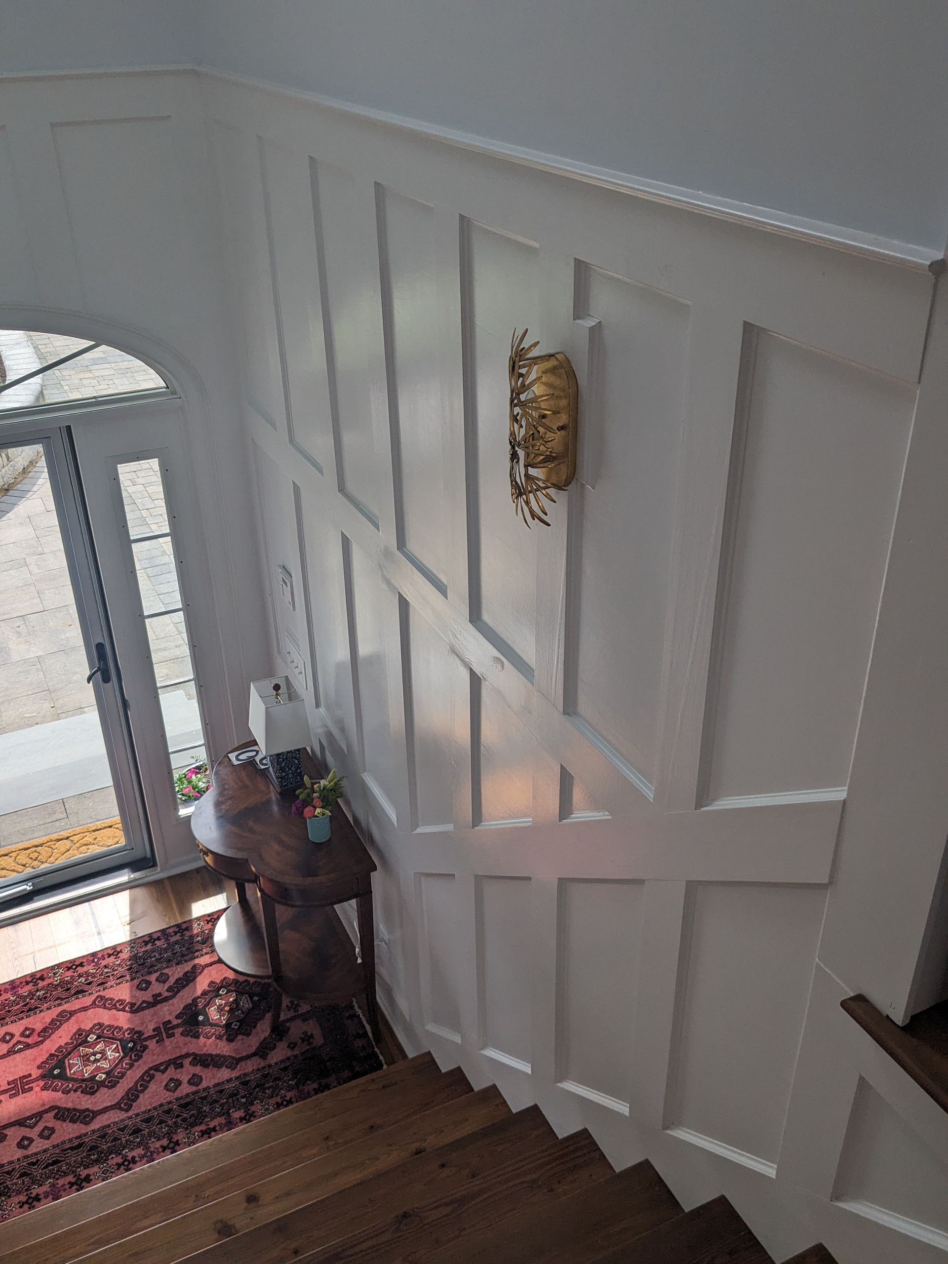 A wooden staircase looking down into an entryway with white wainscoting, a gold wall sconce, and a small dark console table.