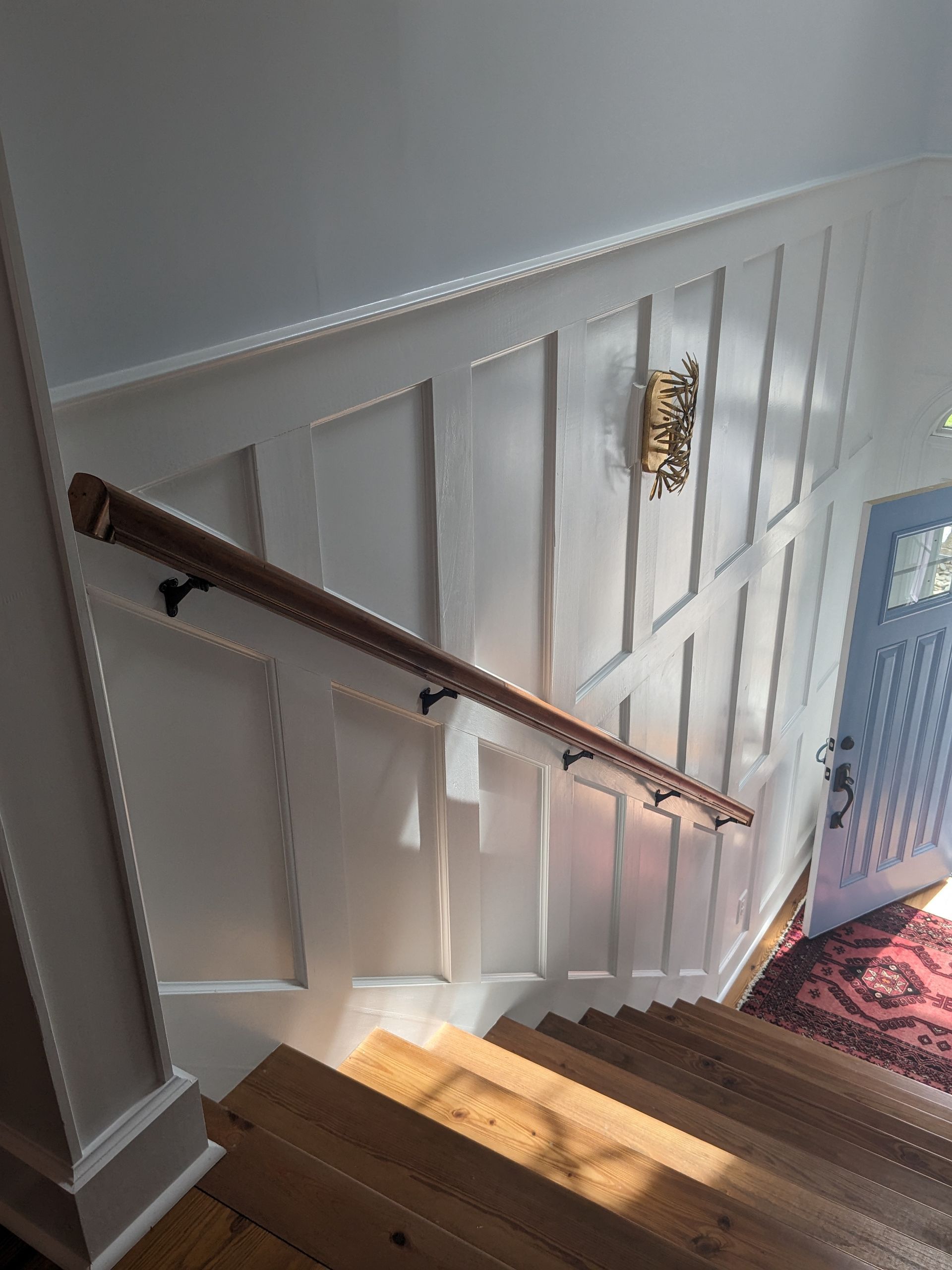 A wooden handrail follows a staircase lined with white wainscoting, leading down to a patterned rug and a light blue door.