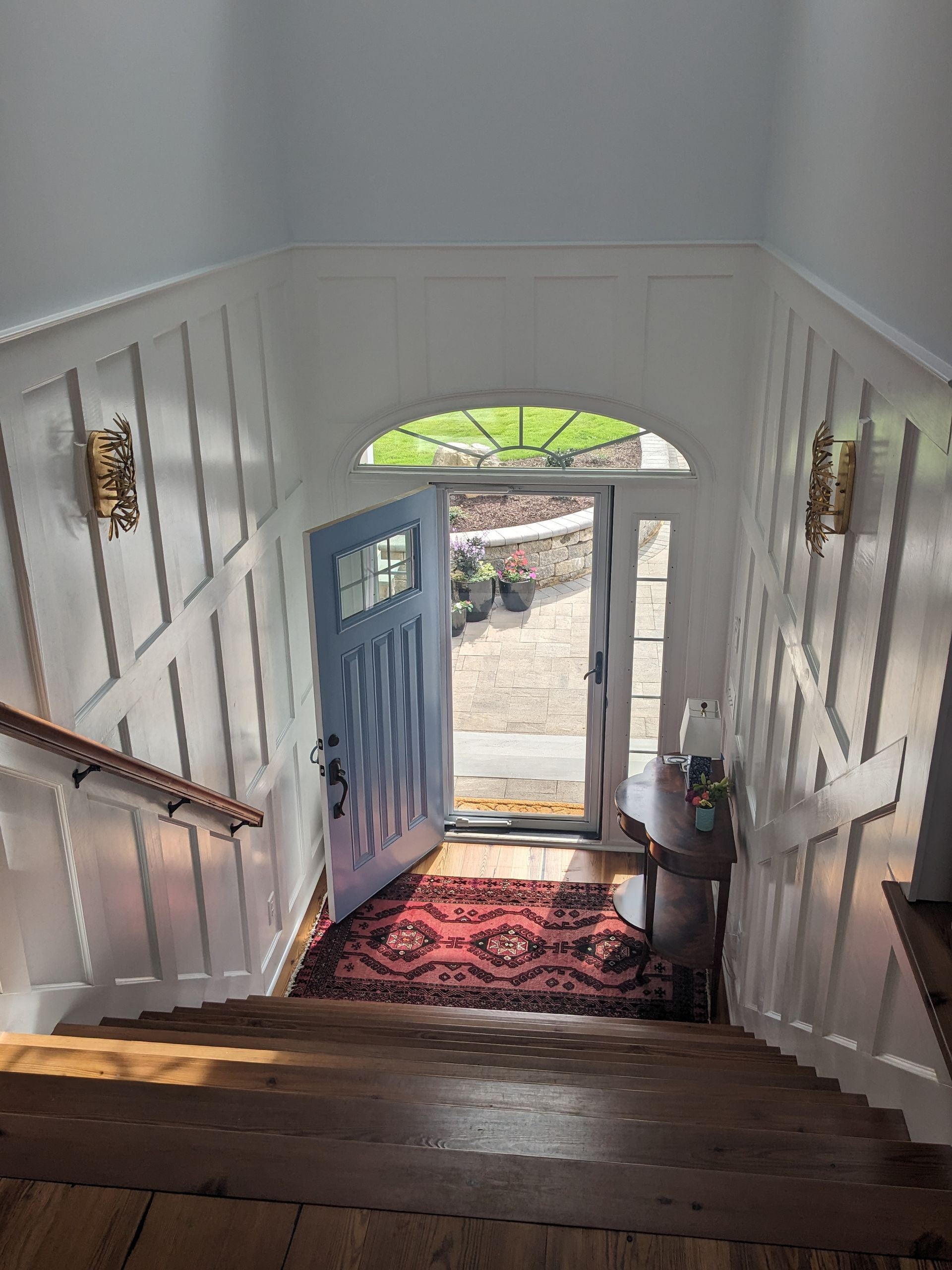 A high-angle view looking down wooden stairs toward a blue front door with a glass transom, white wainscoting, and a rug.