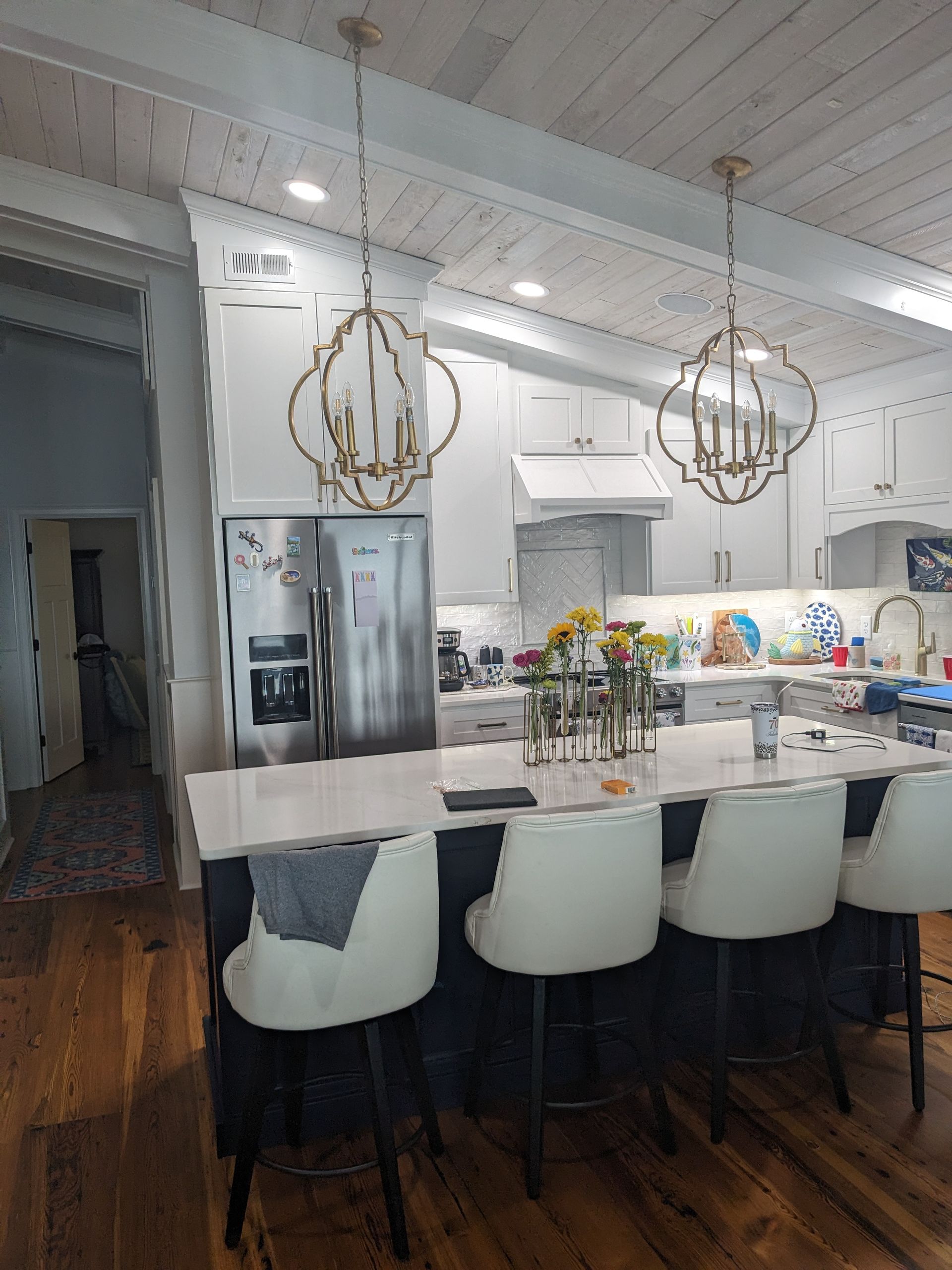 A modern kitchen with a white island and three stools, pendant lights, wooden floors, and white cabinetry.