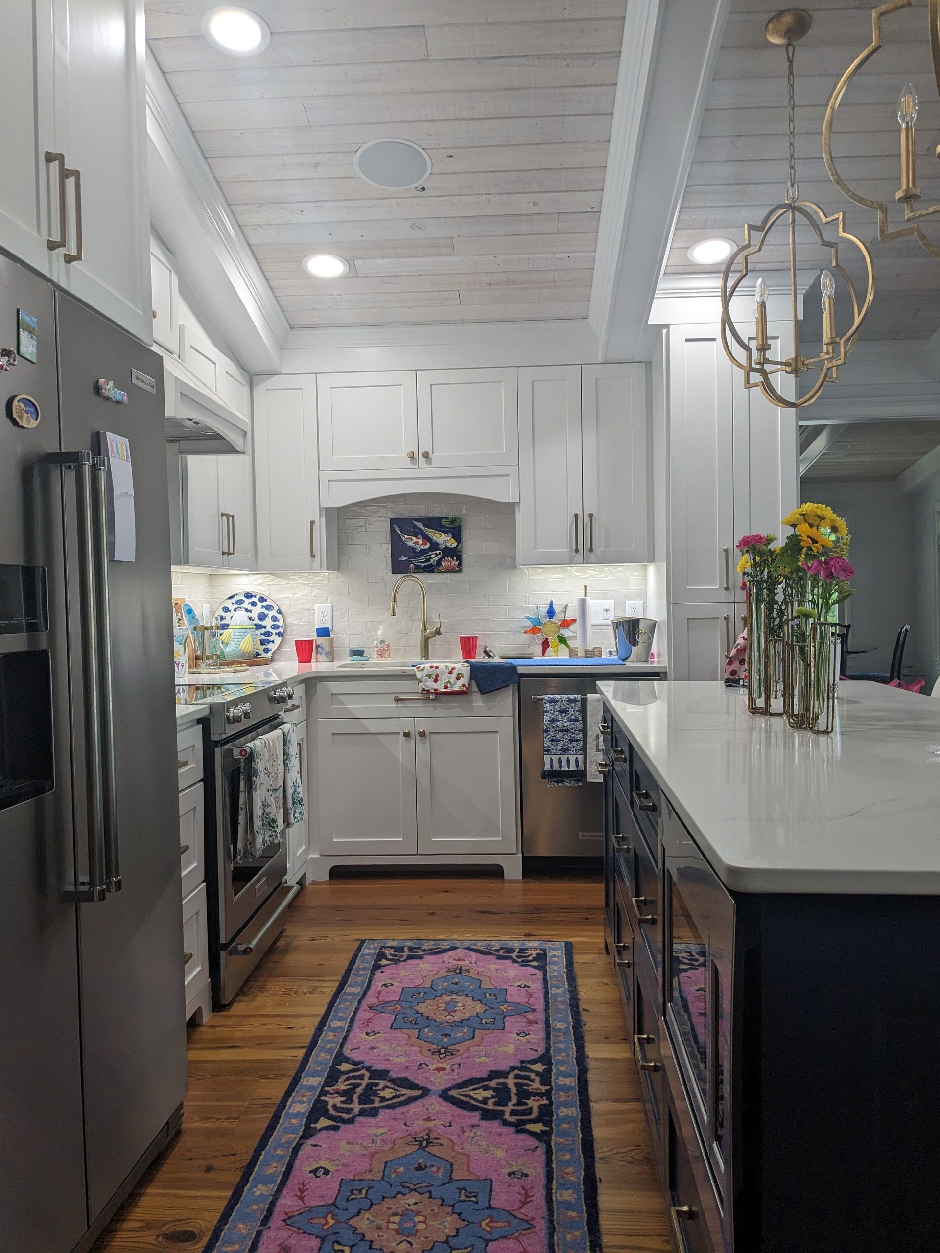 A bright kitchen with white cabinetry, a white shiplap ceiling, a large kitchen island, and a vibrant pink patterned rug.