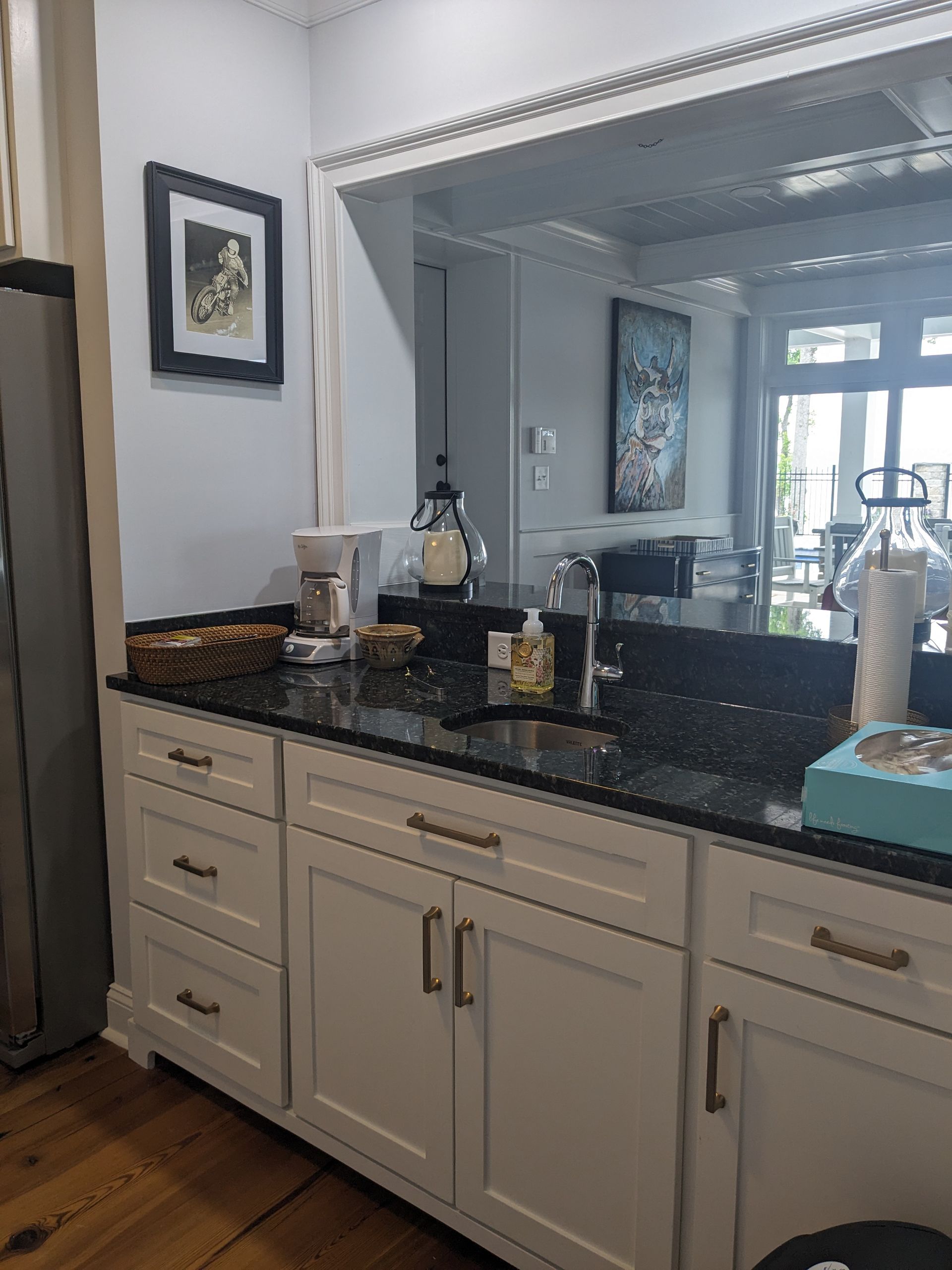 A kitchen wet bar with white cabinets, black granite countertops, a coffee maker, sink, and a large mirror.