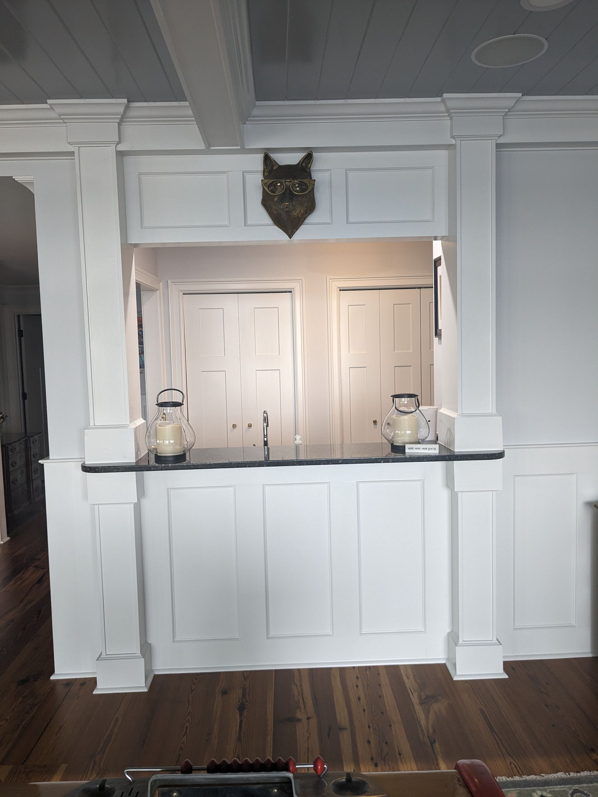 A white home bar with paneled cabinetry, columns, a black countertop, and an animal-head wall decor piece.