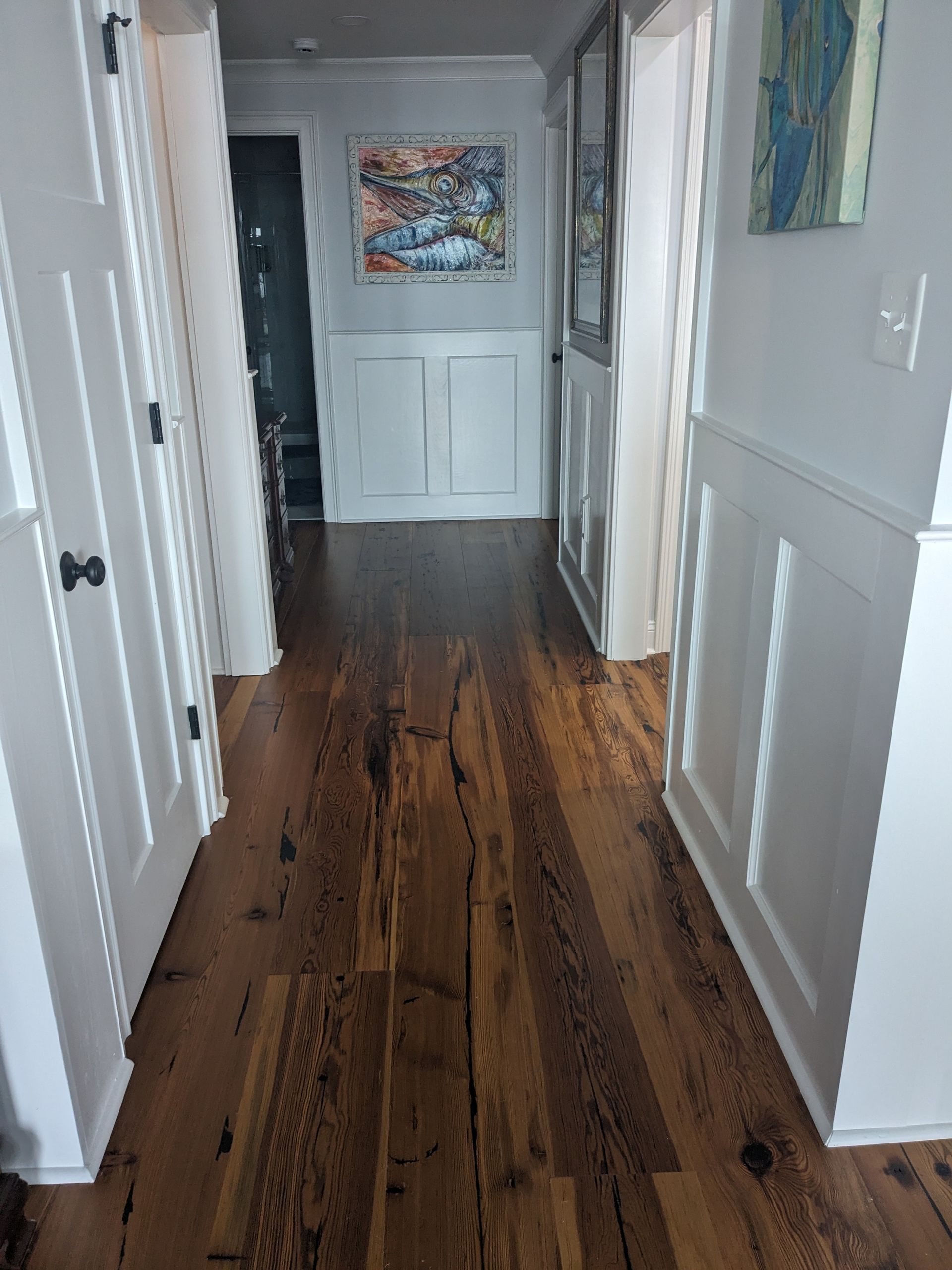 A hallway with dark wood floors, white wainscoting, and white doors leading to rooms with art hanging on the walls.