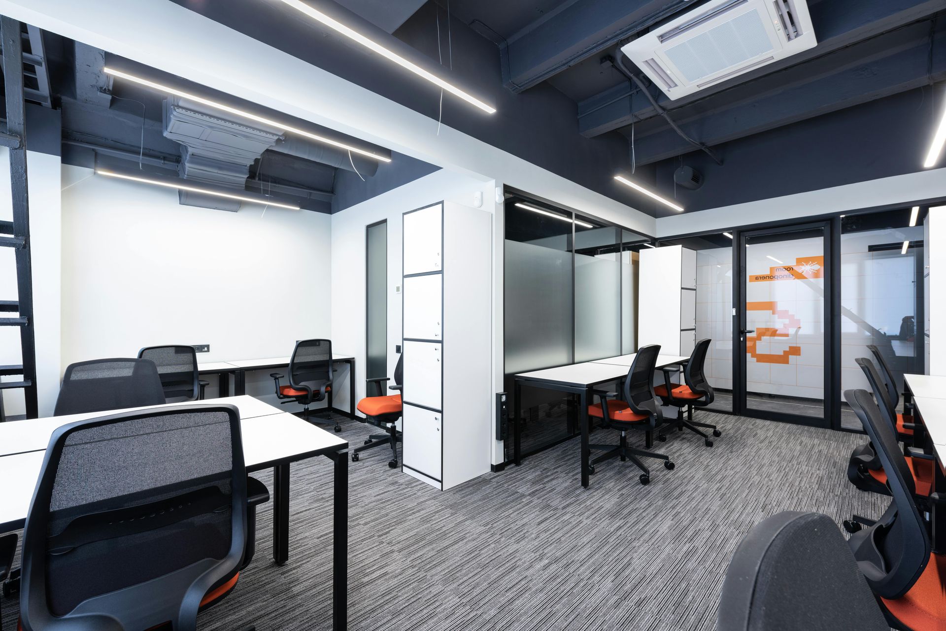 Modern office space featuring white desks with orange-accented chairs, a dark ceiling, and patterned gray carpet flooring.