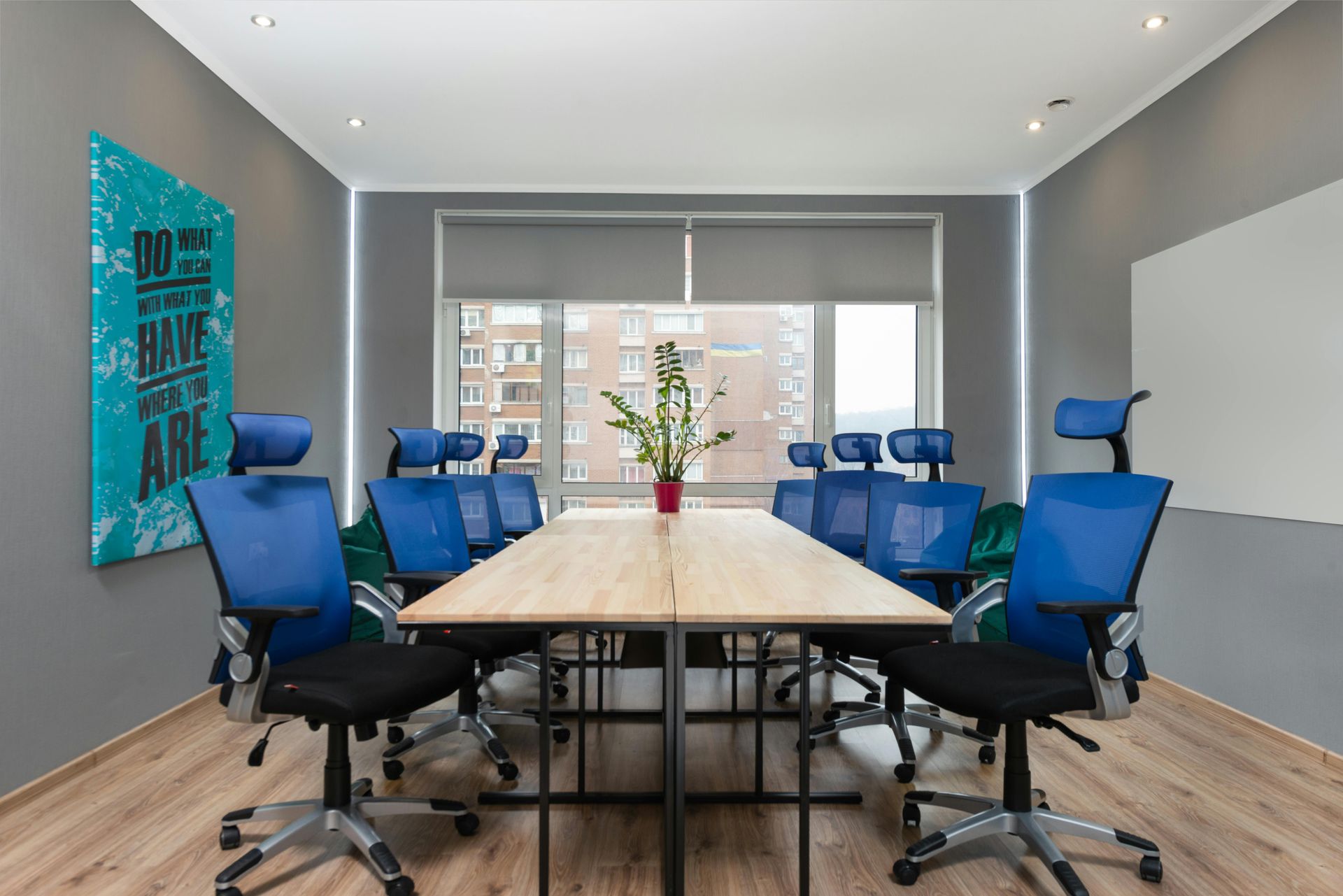 A modern office meeting room with a long wooden table surrounded by blue mesh chairs, a window, and gray walls.