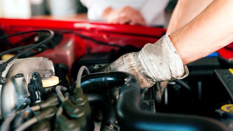 Mechanic's gloved hands working on a car engine, focusing on components. Red car hood visible.