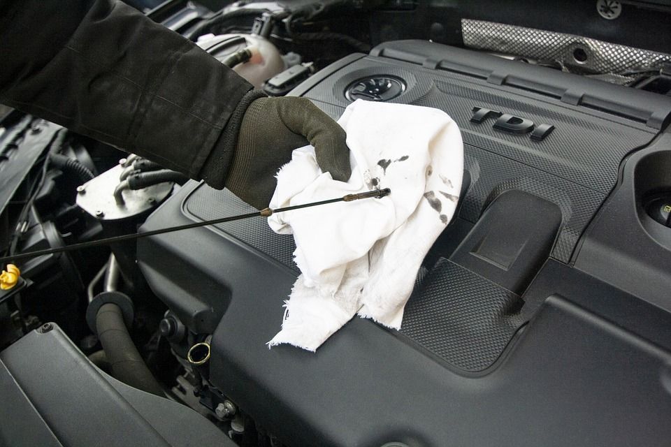 Gloved hand wiping a car's oil dipstick with a cloth, next to a black engine.
