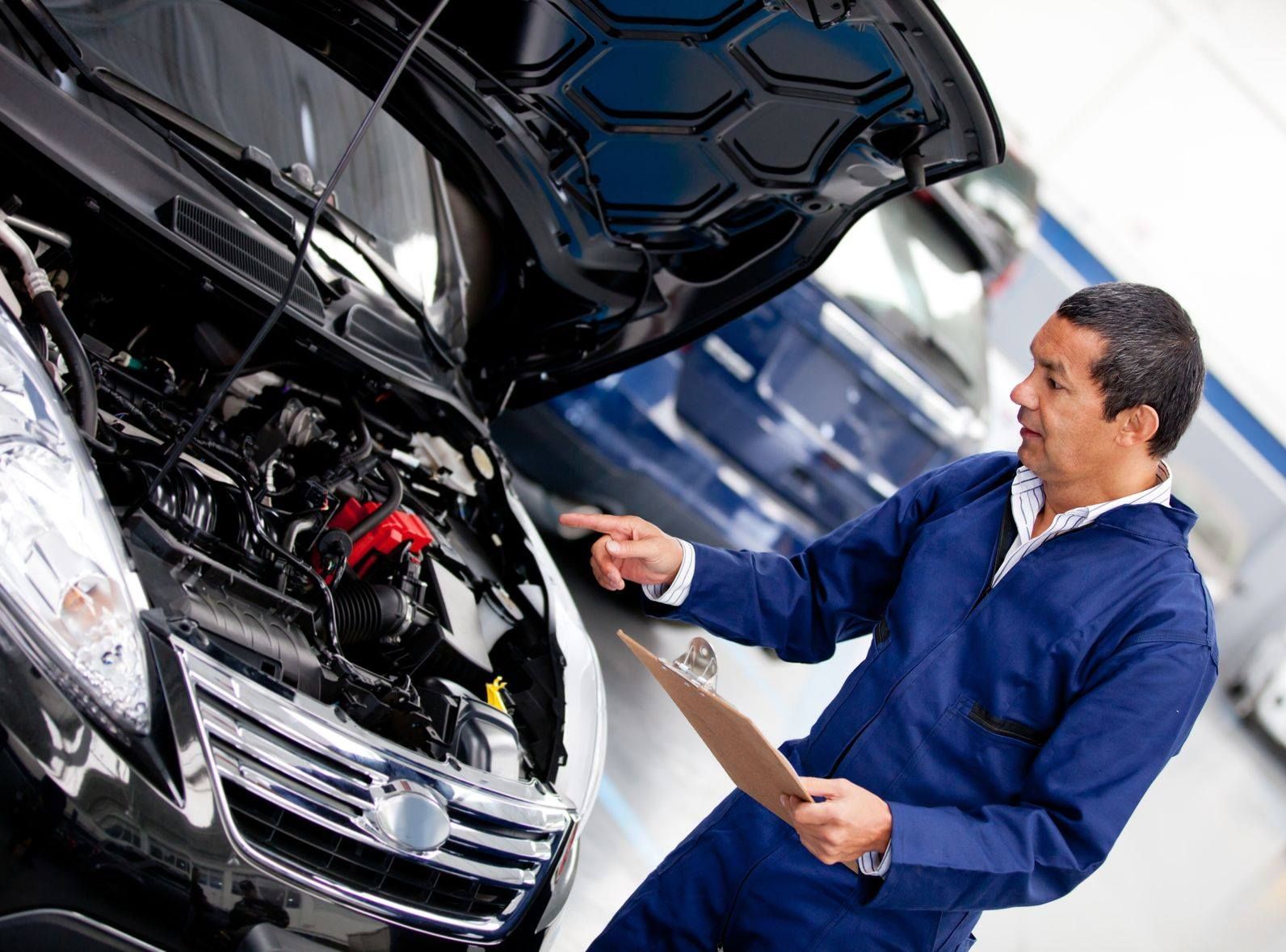 Mechanic in blue jumpsuit examining car engine with clipboard in a garage.