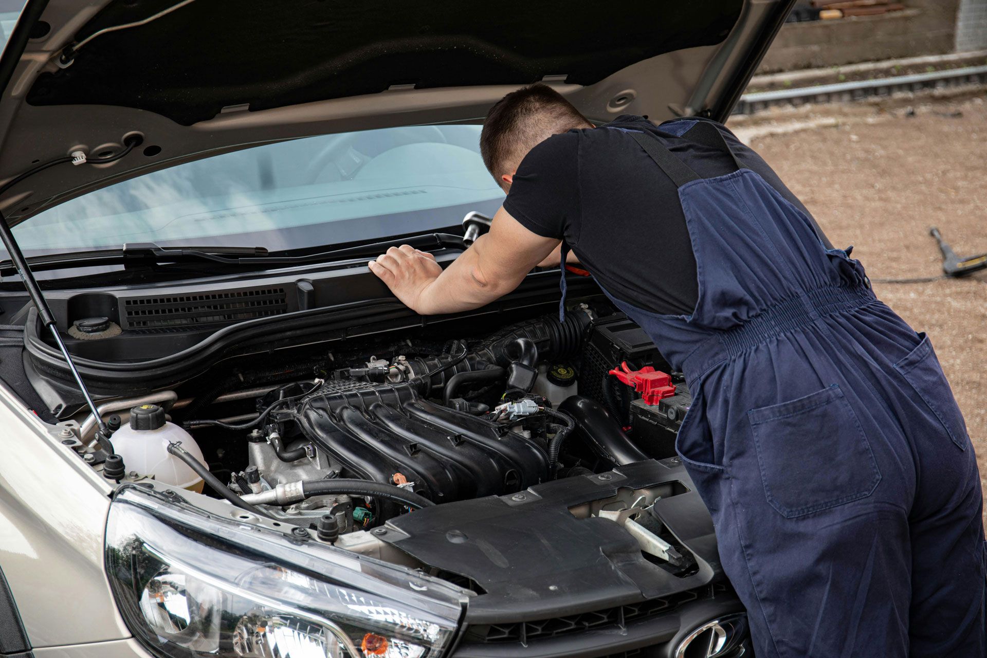 Mechanic in blue overalls uses a diagnostic tool on an orange car with its hood open in a garage.