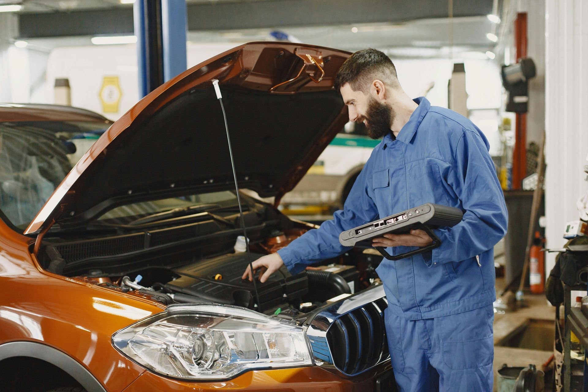 Mechanic in blue uniform inspects an orange car engine with a diagnostic tool in a garage.