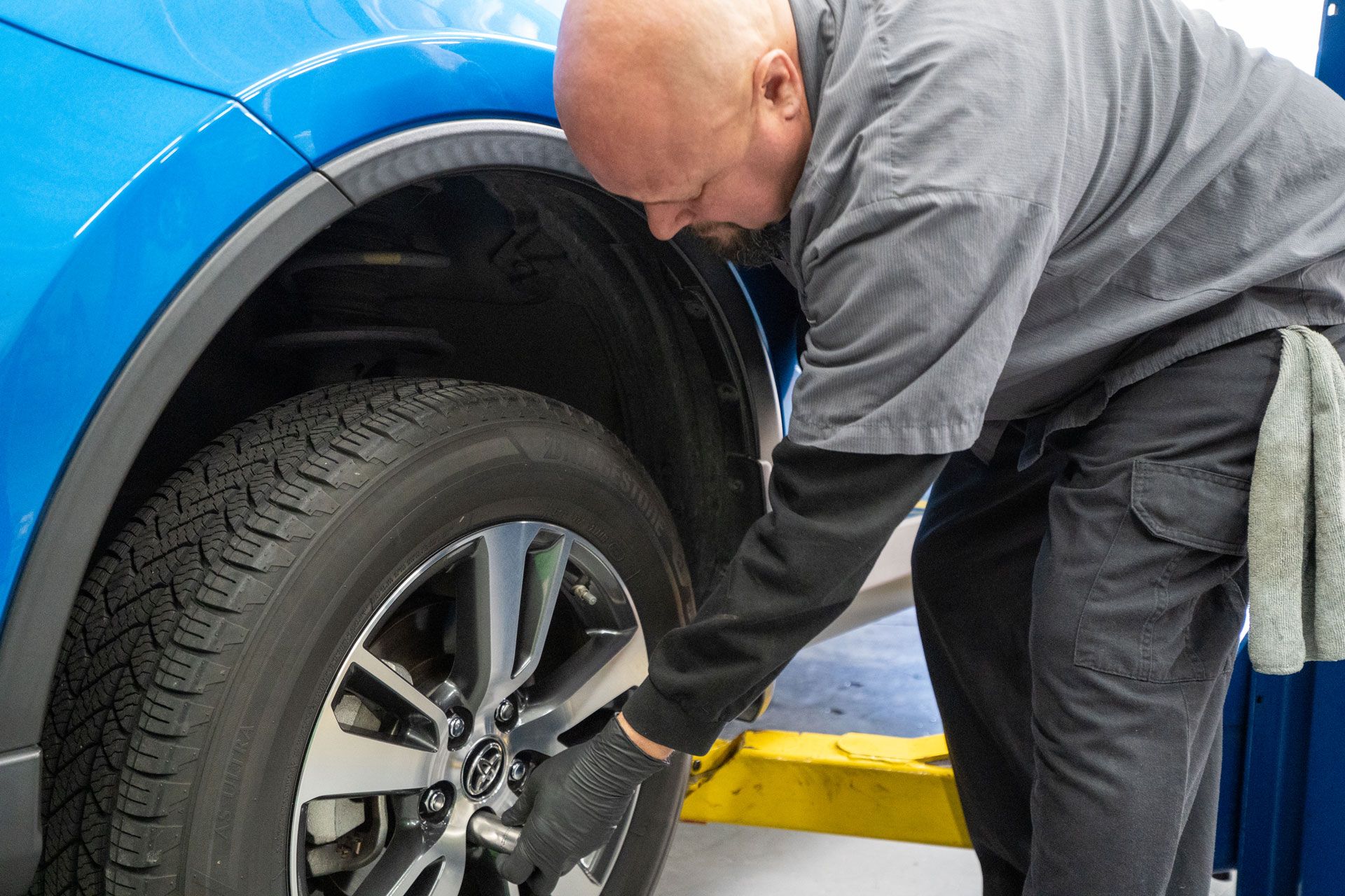 Mechanic works on blue car tire; he's wearing black gloves and gray work clothes. Garage setting.