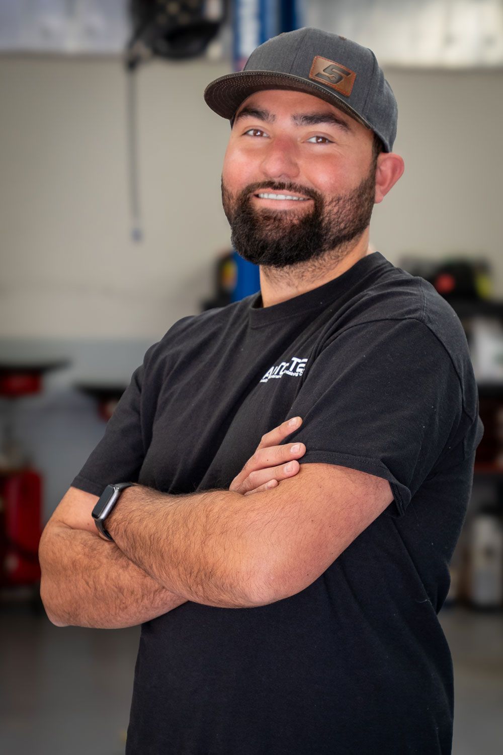 Man in a cap and black shirt with arms crossed, smiling in a garage setting.