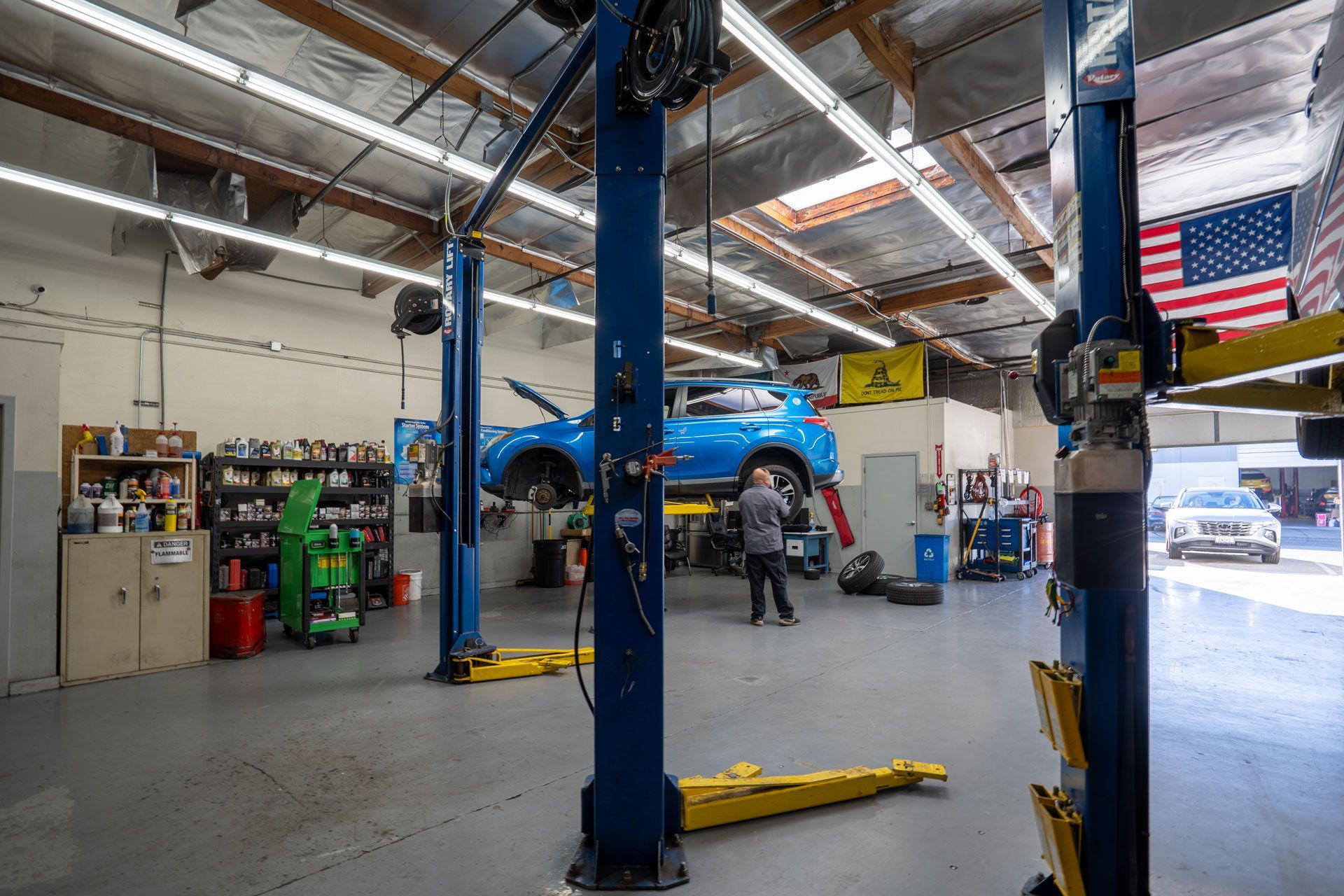 Auto repair shop interior. A blue car is raised. A mechanic works. American flag is on a wall.