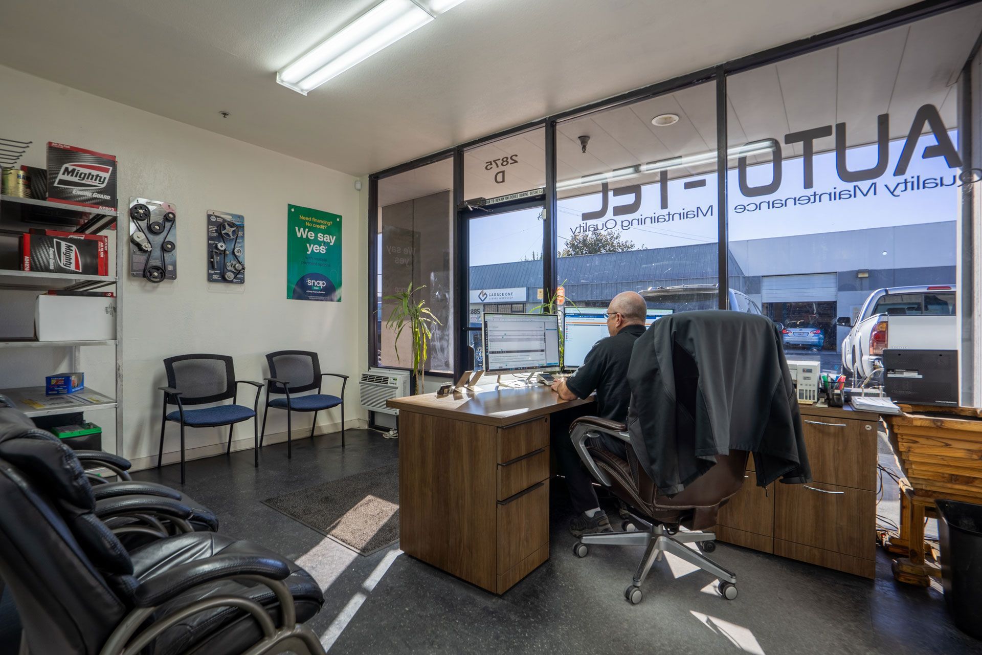 An office interior with a person at a desk. There are chairs, a window, and automotive parts on the wall.