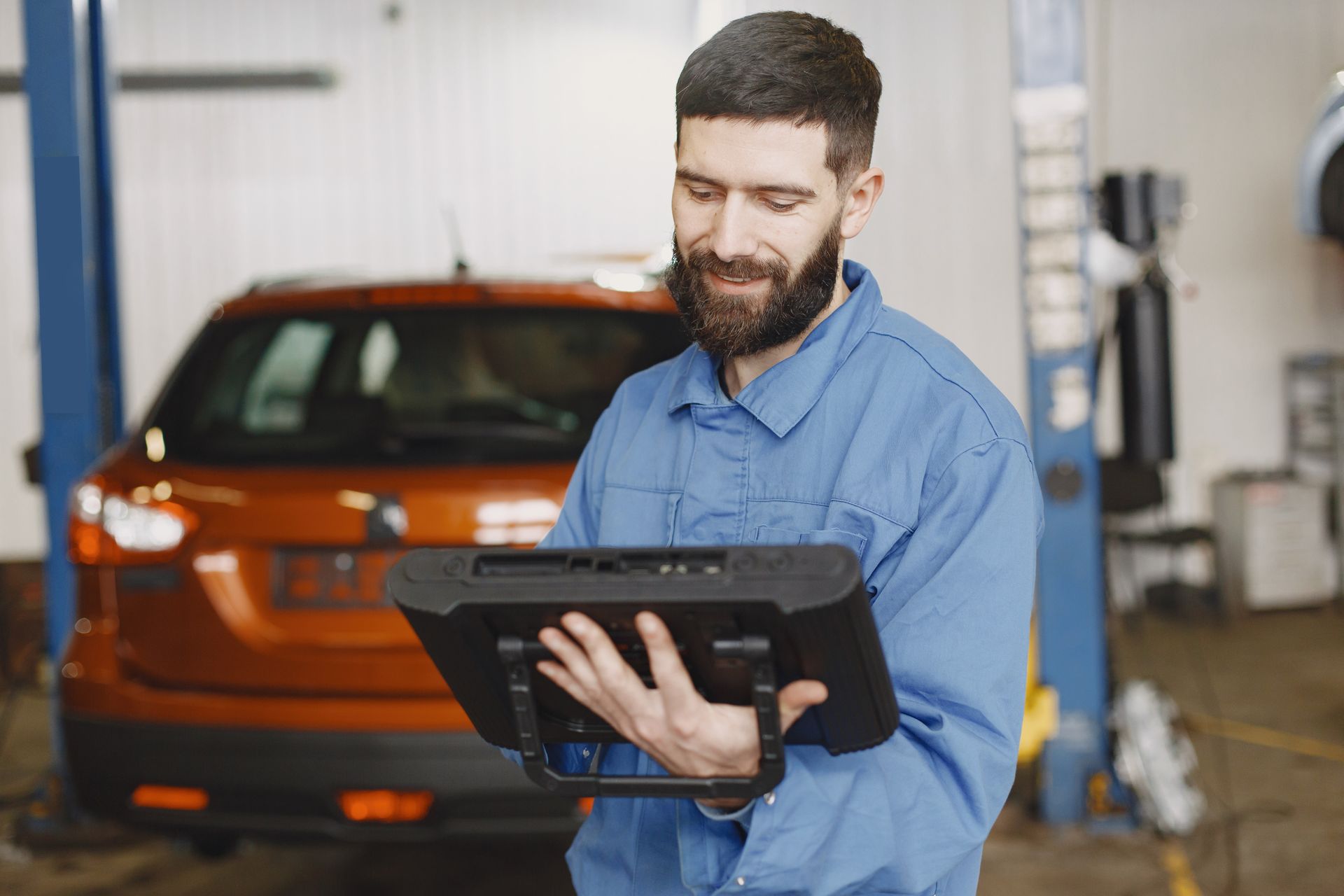 Mechanic in blue overalls uses a diagnostic tablet in a garage near an orange car.