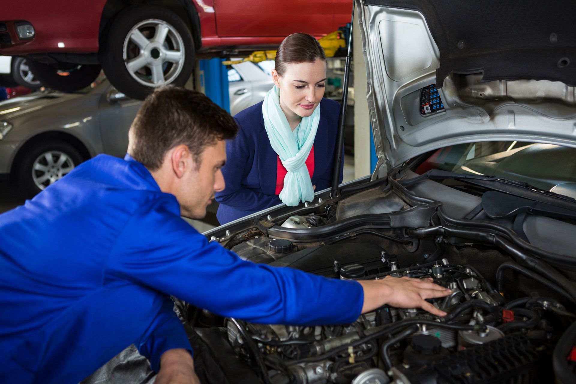 Mechanic in blue uniform pointing at car engine, explaining to a woman in a blazer at a garage.