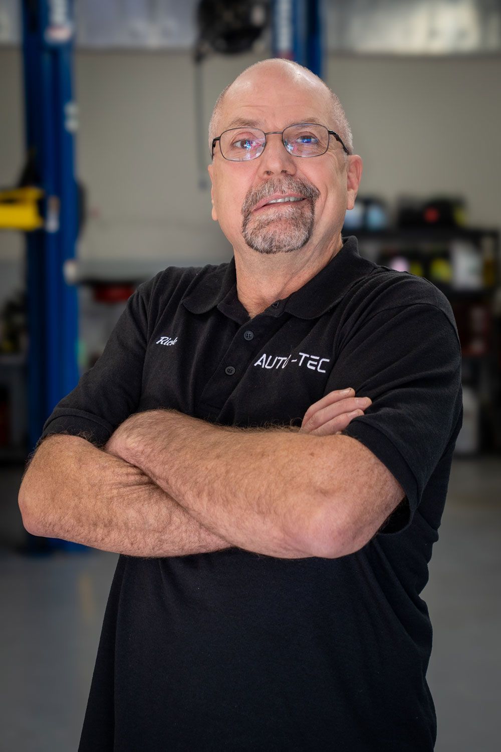Man with glasses and a goatee in a black shirt with logo; arms crossed, smiles in garage setting.
