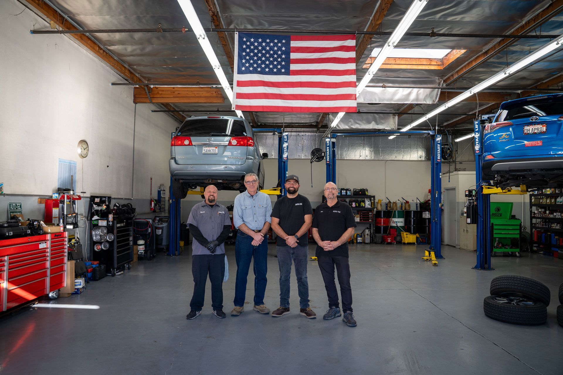 Four men in a garage with an American flag. A car is raised on a lift. Shop tools visible.