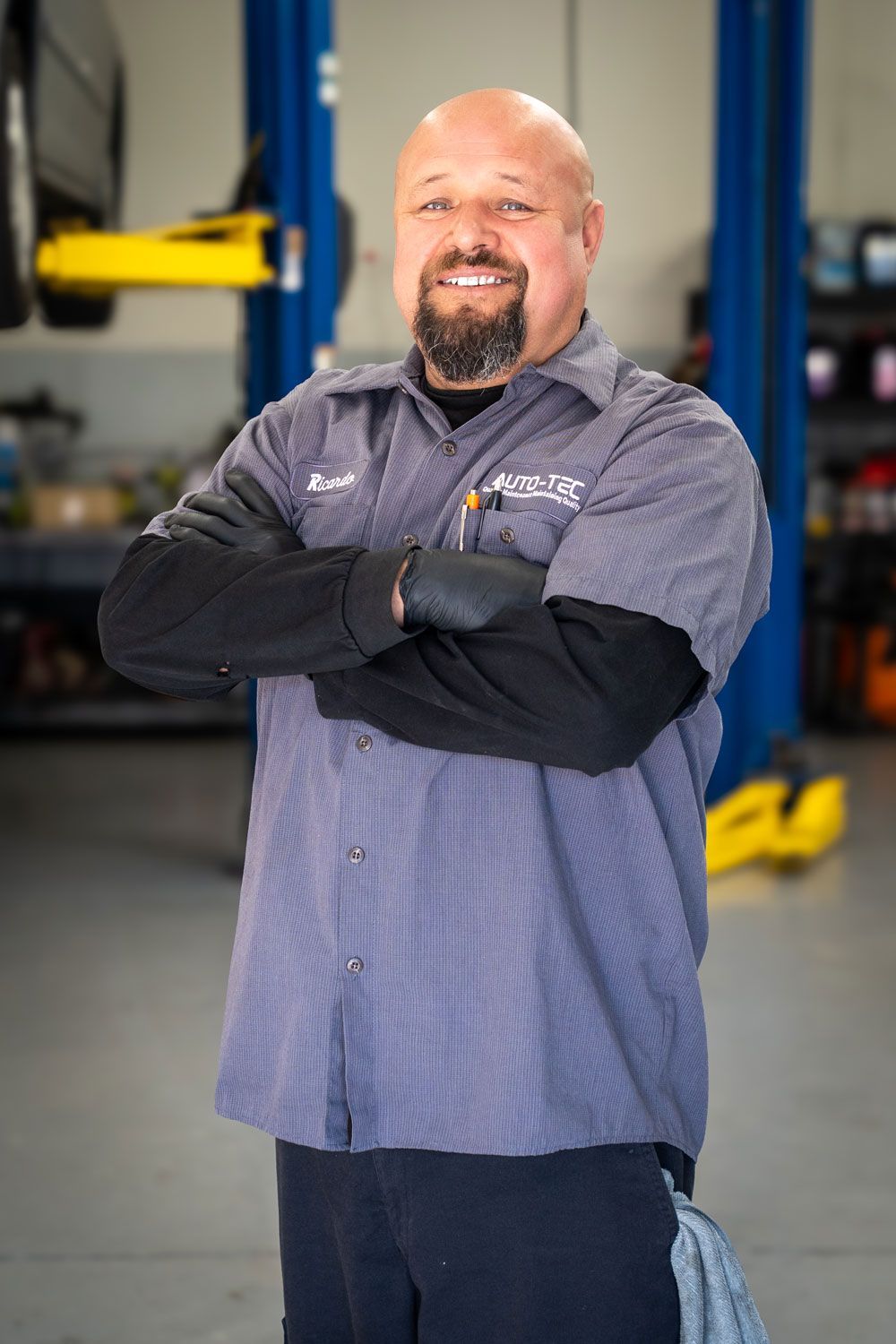 Mechanic in blue work shirt, arms crossed, smiling in a garage with car lift.