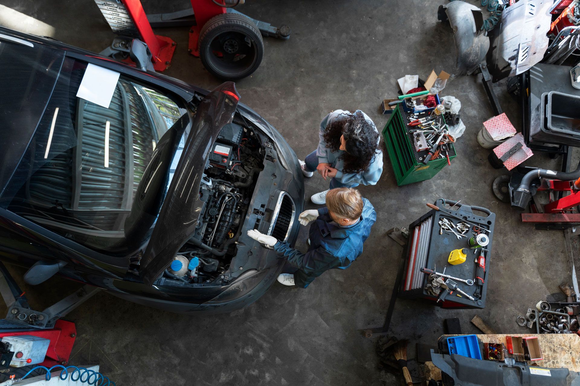 People examining car engine in auto repair shop. Tools and parts are scattered around.