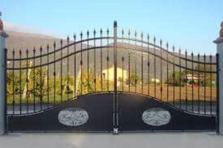 Un cancello in ferro battuto nero è aperto su un campo con una casa sullo sfondo.