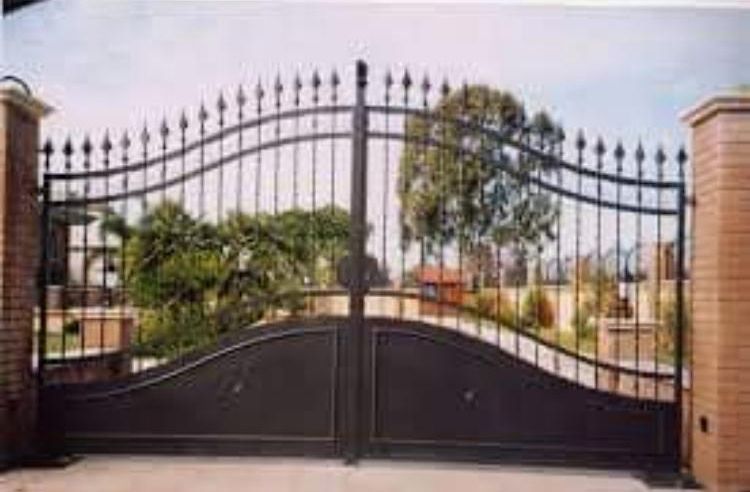 Un cancello in ferro battuto nero è aperto su un vialetto con alberi sullo sfondo.