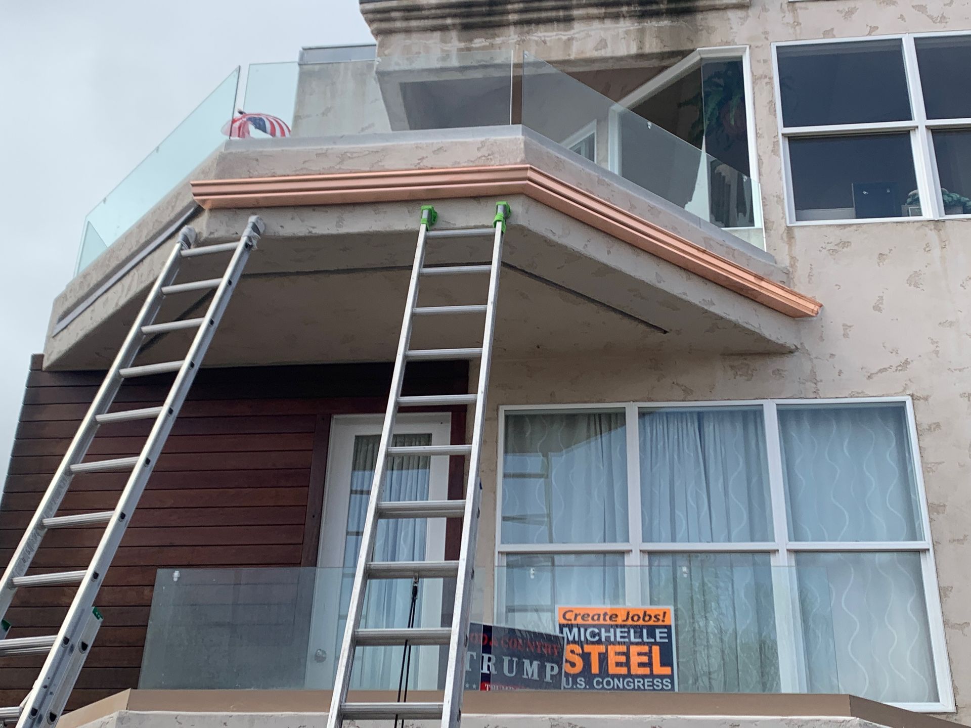 Two ladders lean against a multi-story building with a rooftop deck and glass railing.