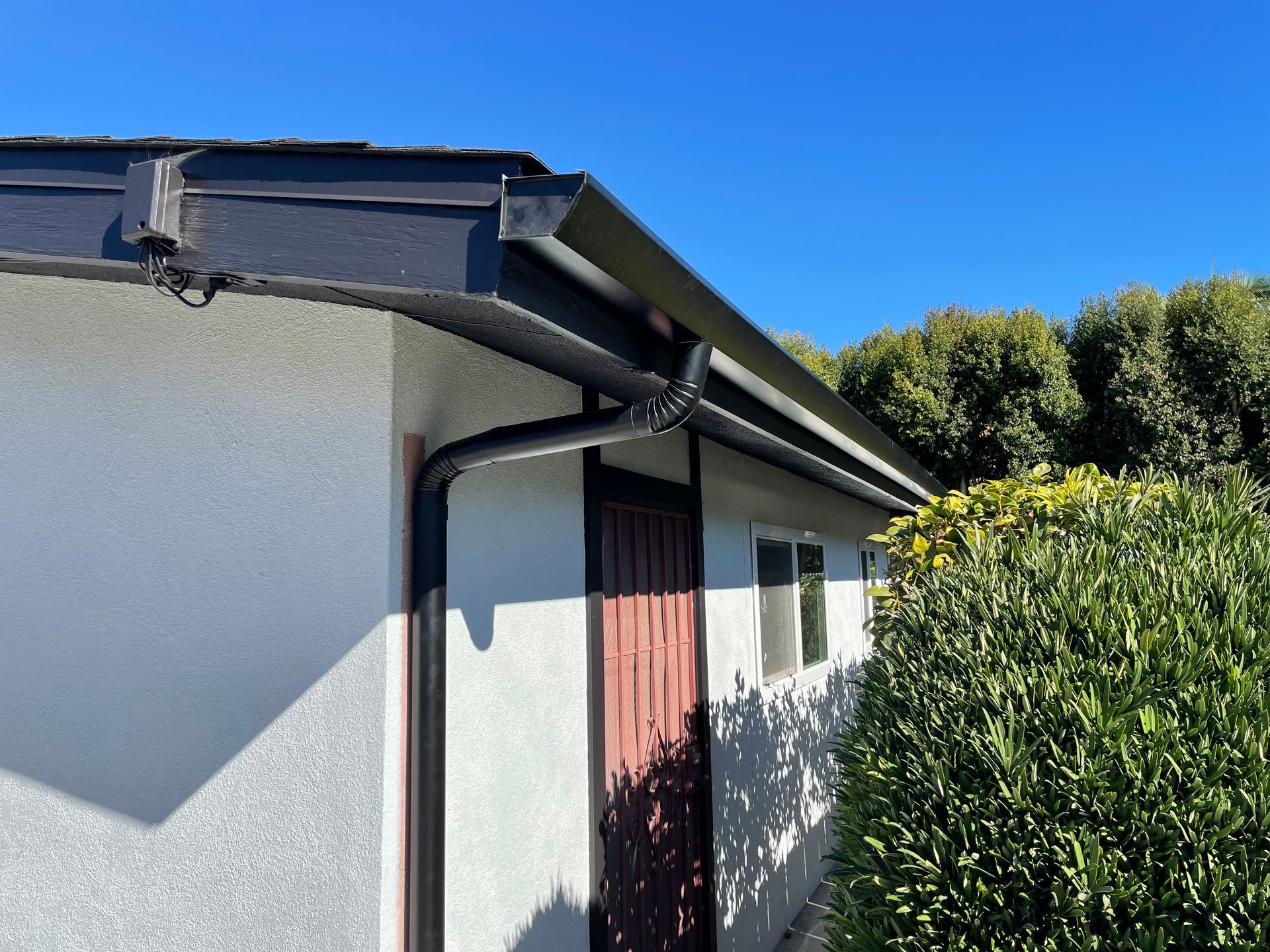Black rain gutters on a white stucco building with a brown door and a clear, blue sky.