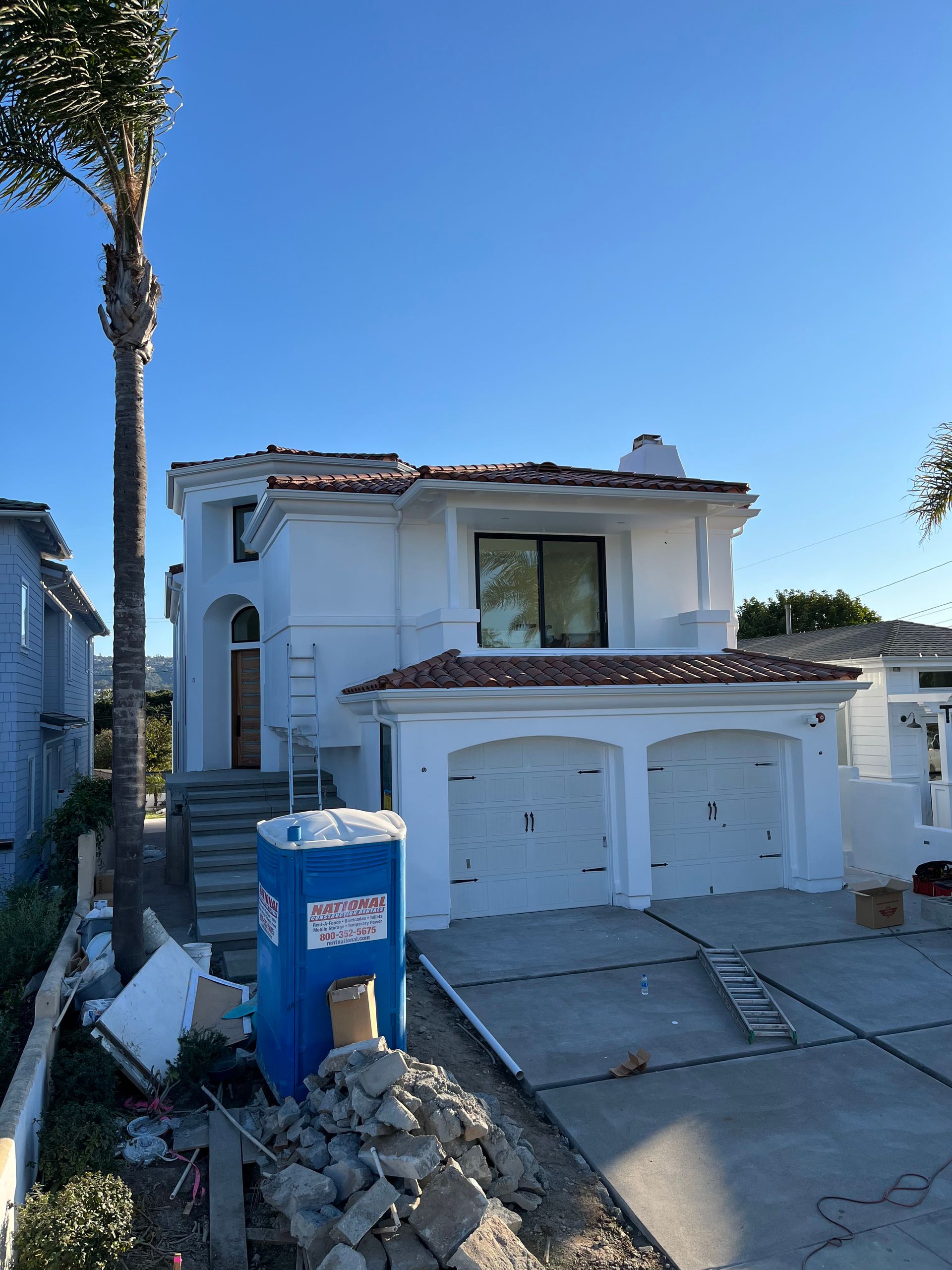 Two-story white house under construction with blue portable toilet and palm tree.