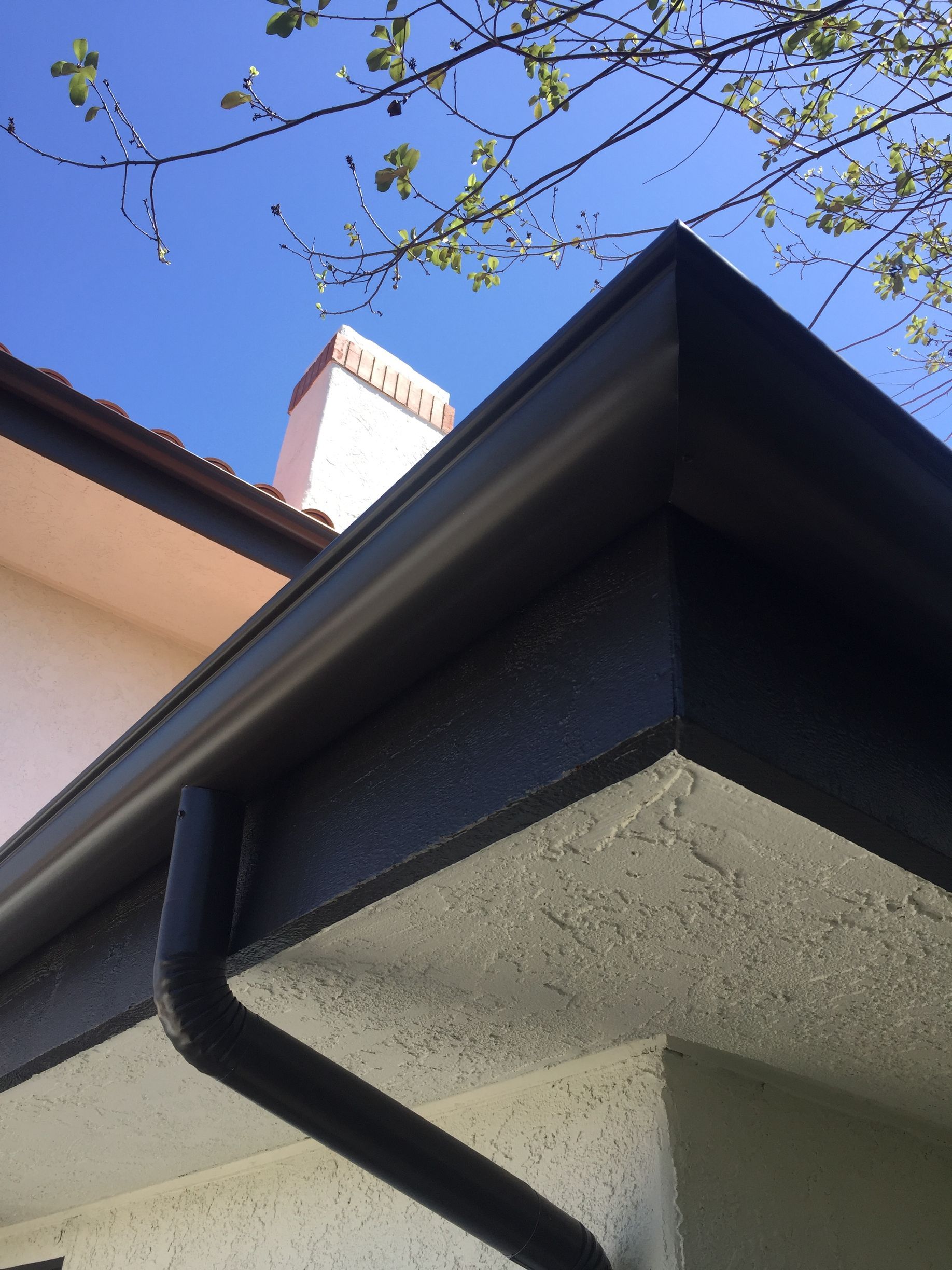 Dark gutter and downspout on a stucco building under a bright blue sky.