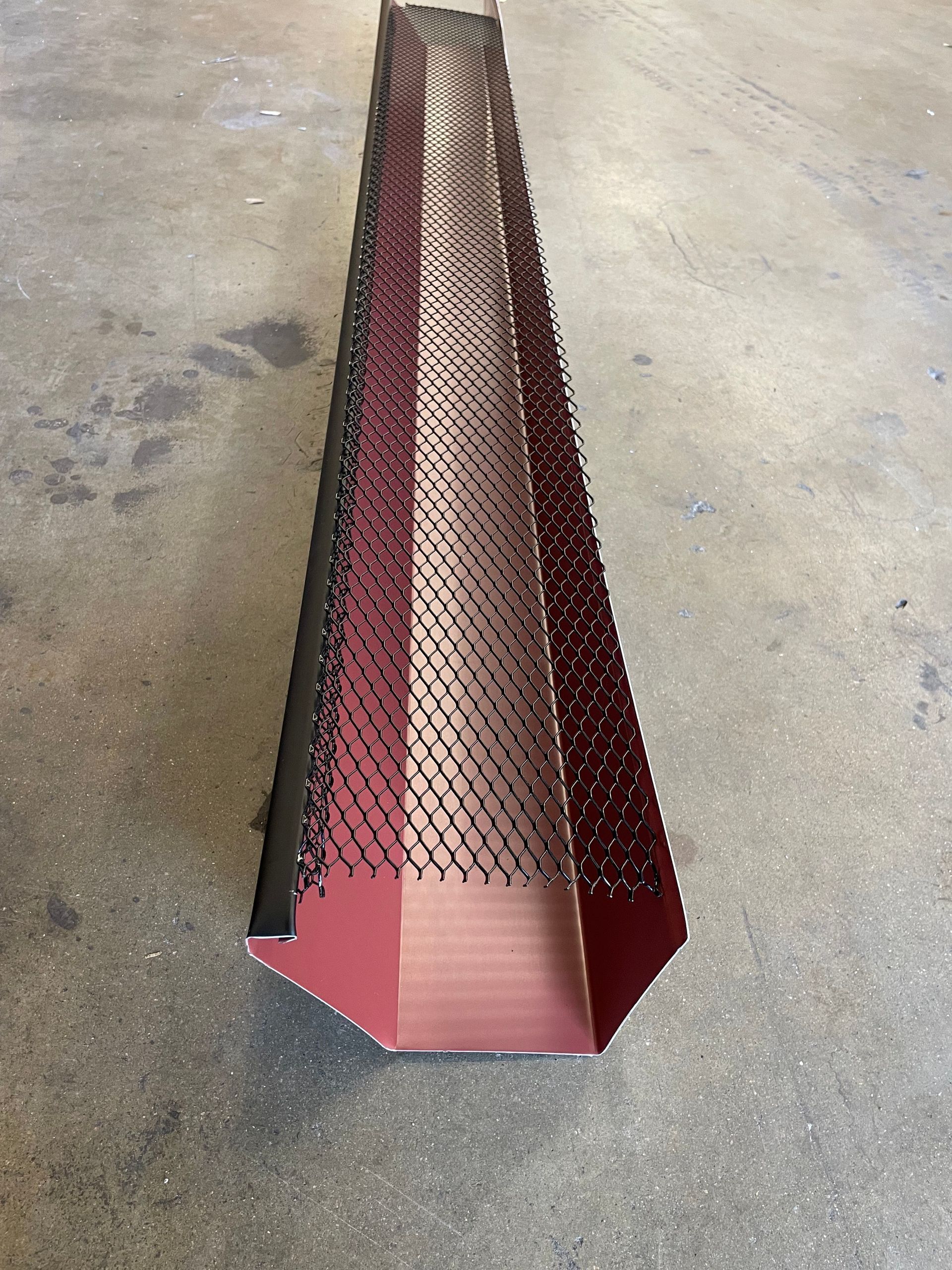 Red and black gutter with wire mesh, resting on a gray concrete surface.