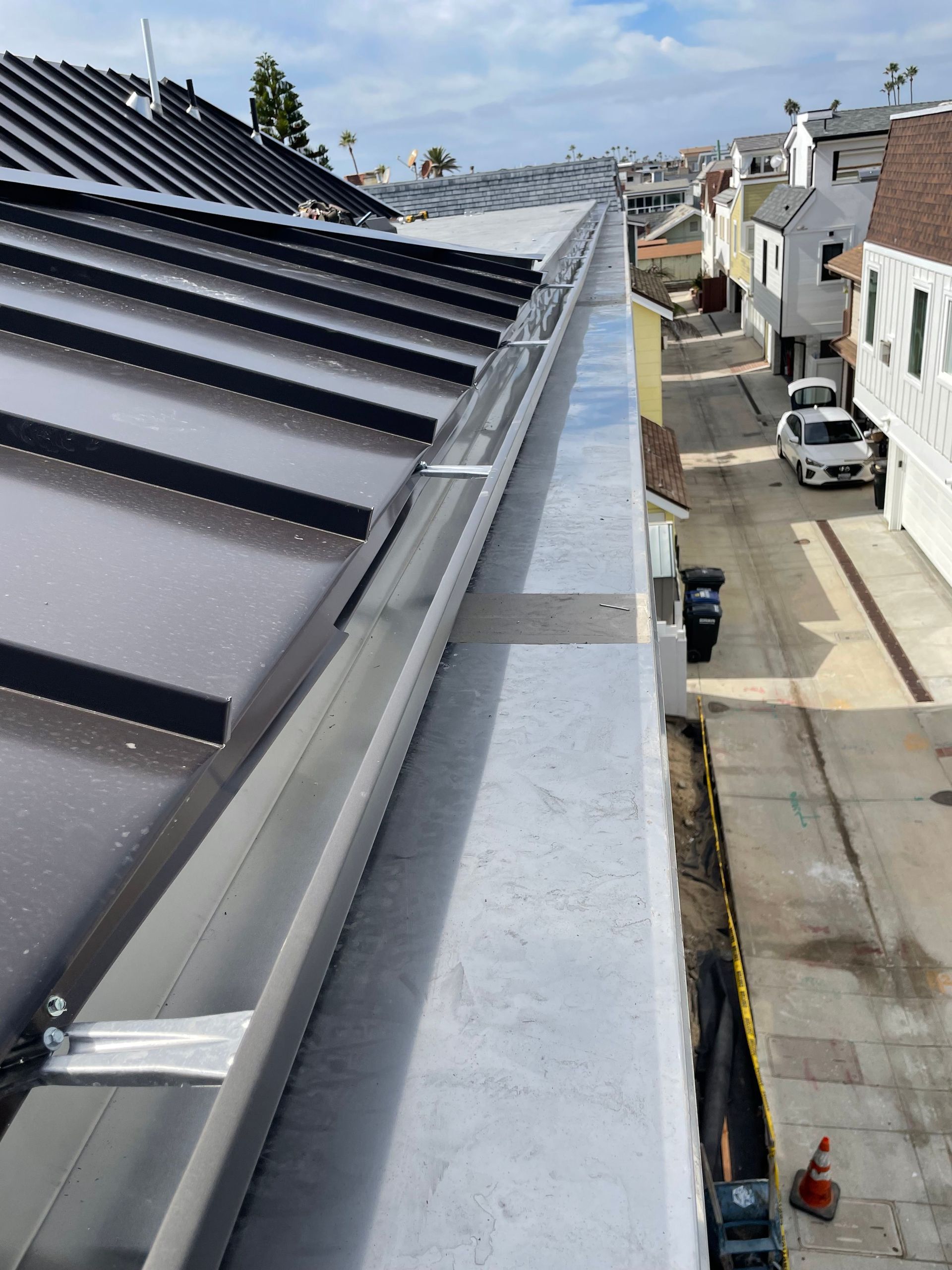 View of a metal roof with a long gutter overlooking a narrow street lined with houses and parked cars.