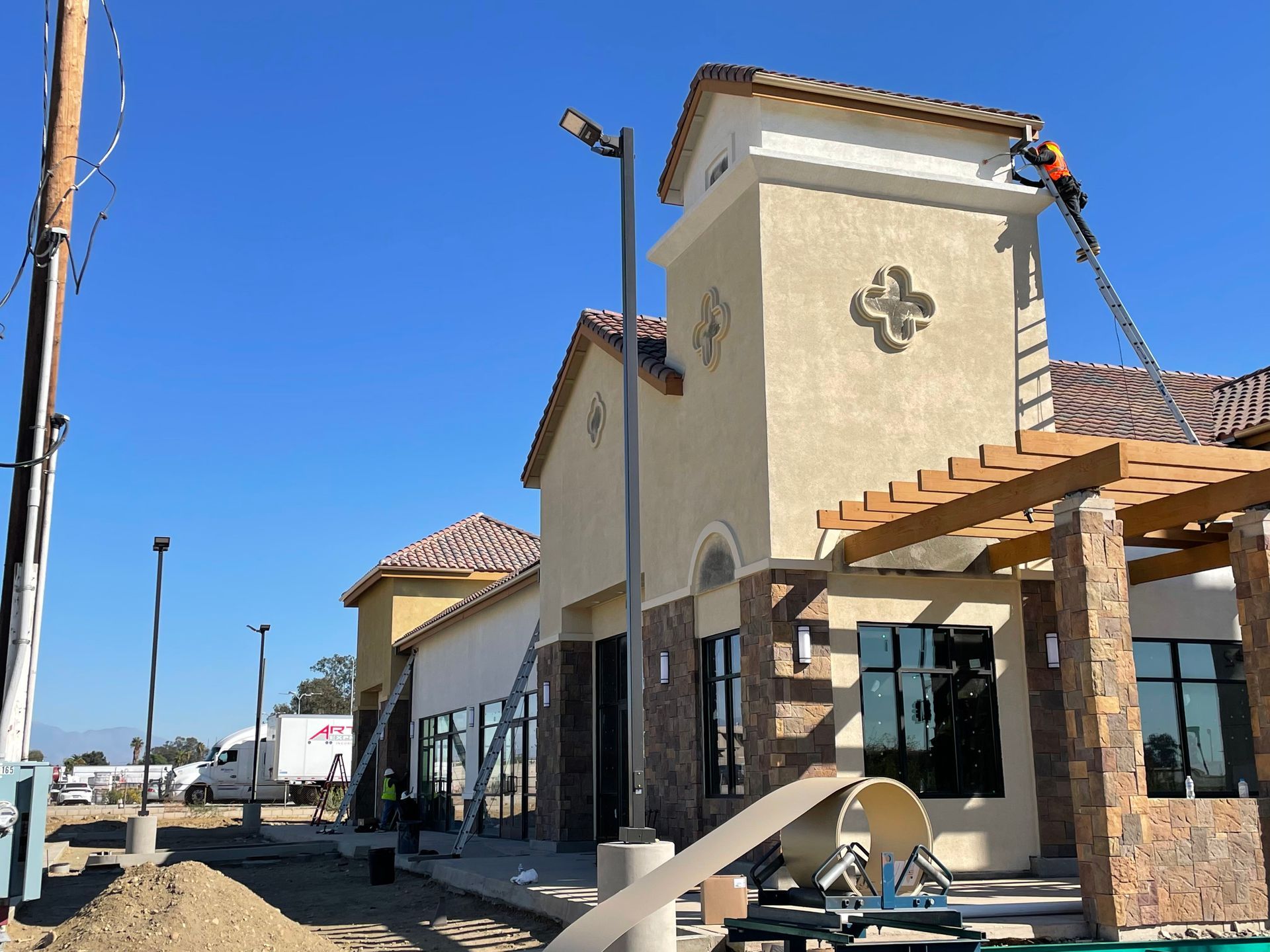 Construction of a tan building with stone accents; a worker on a ladder is working on the roof.