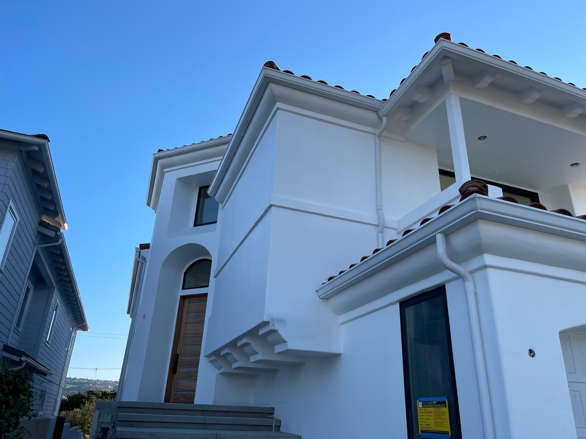 White stucco house with tile roof, next to a grey house, against a clear blue sky.
