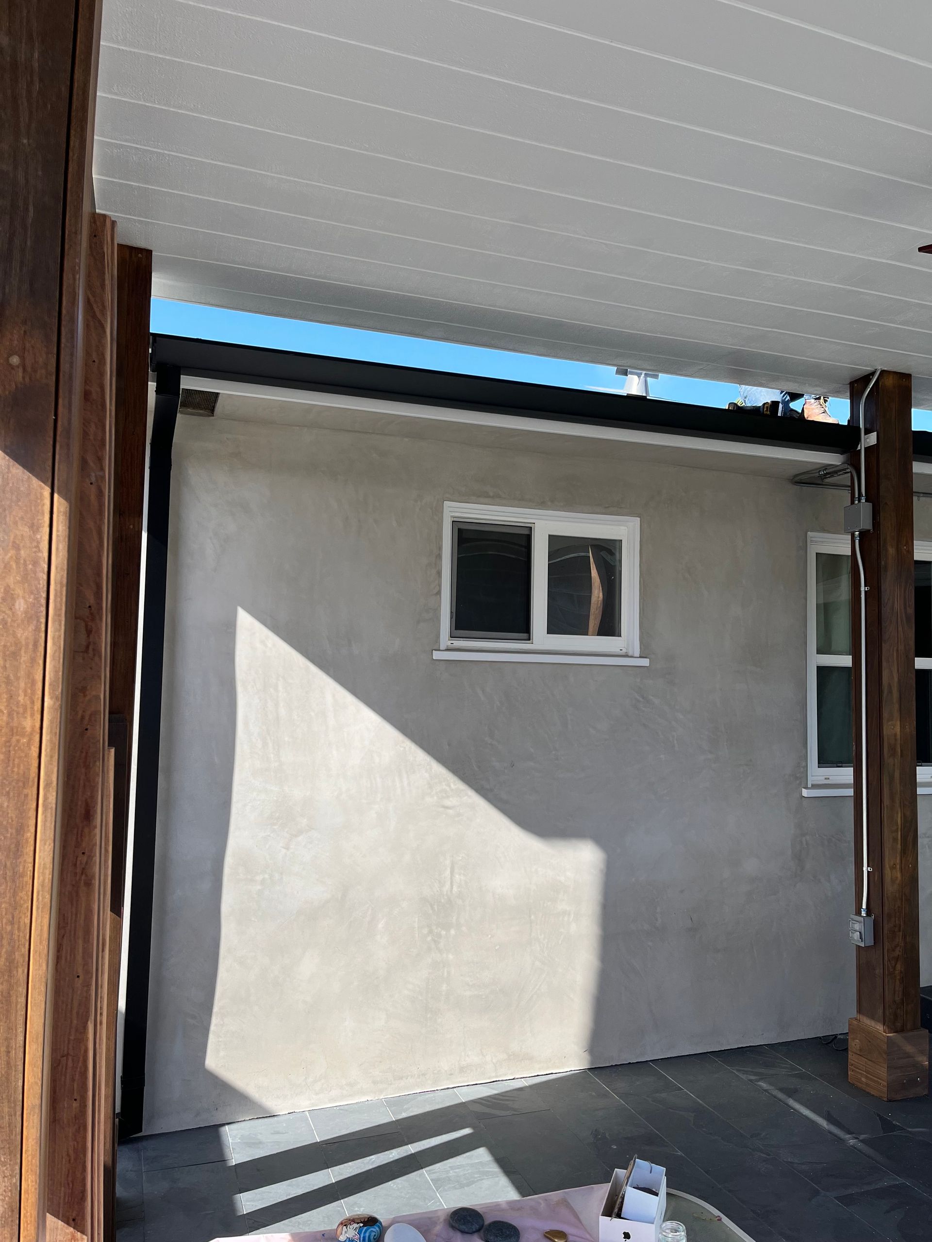 A stucco wall with a window and wooden support beams, shaded by a light gray canopy.