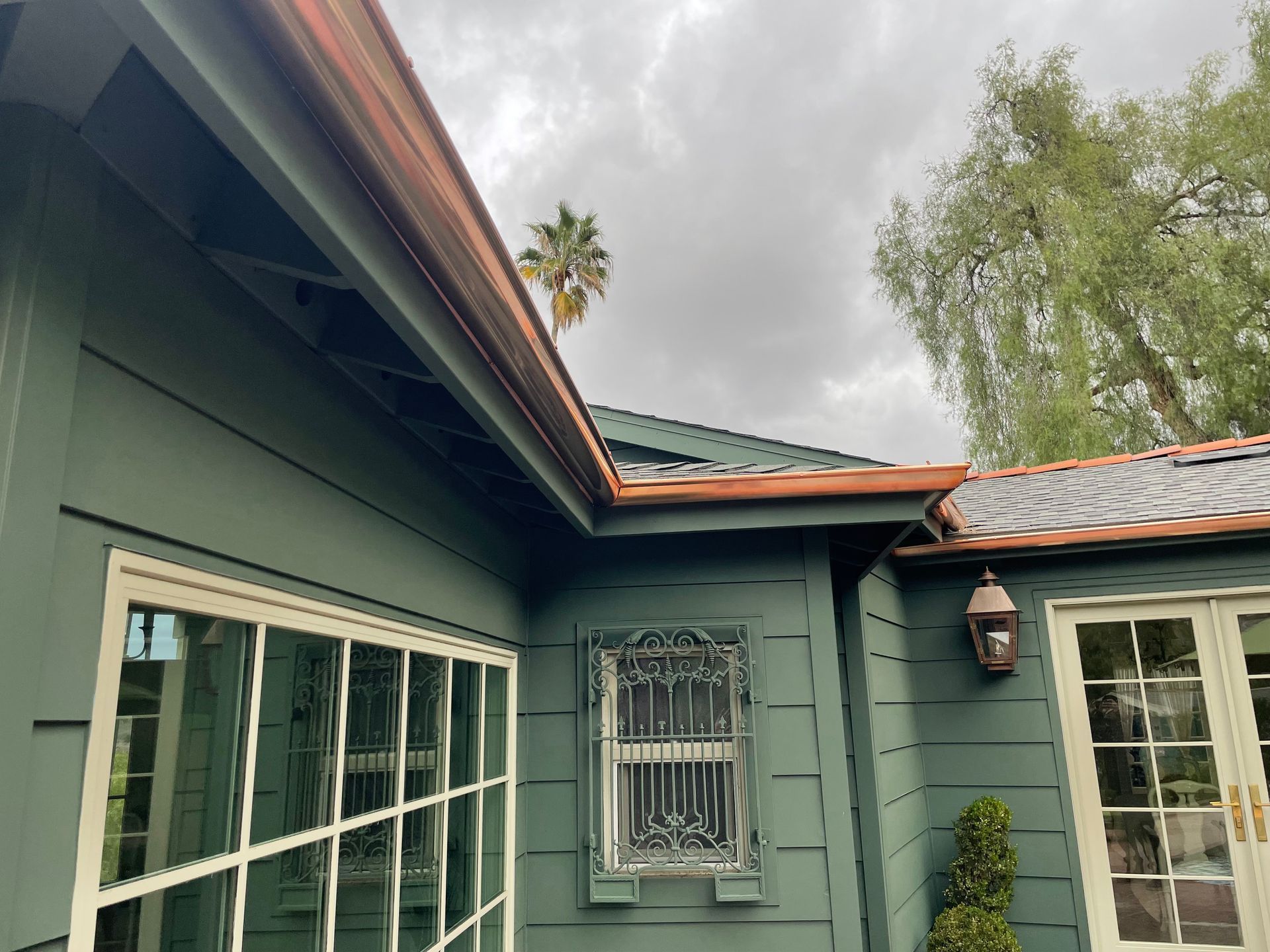 Green house exterior with copper gutters, white-framed windows, and a cloudy sky.